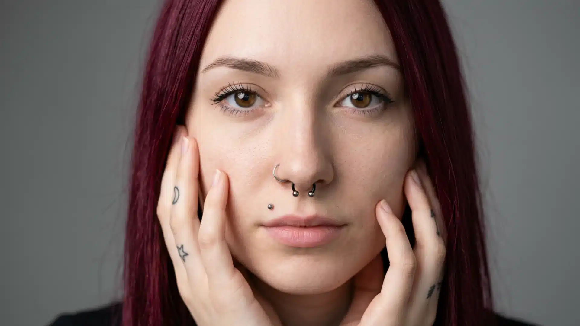 Portrait of a person with maroon hair, a septum ring, nose ring, and lip piercing framing their face with tattooed hands