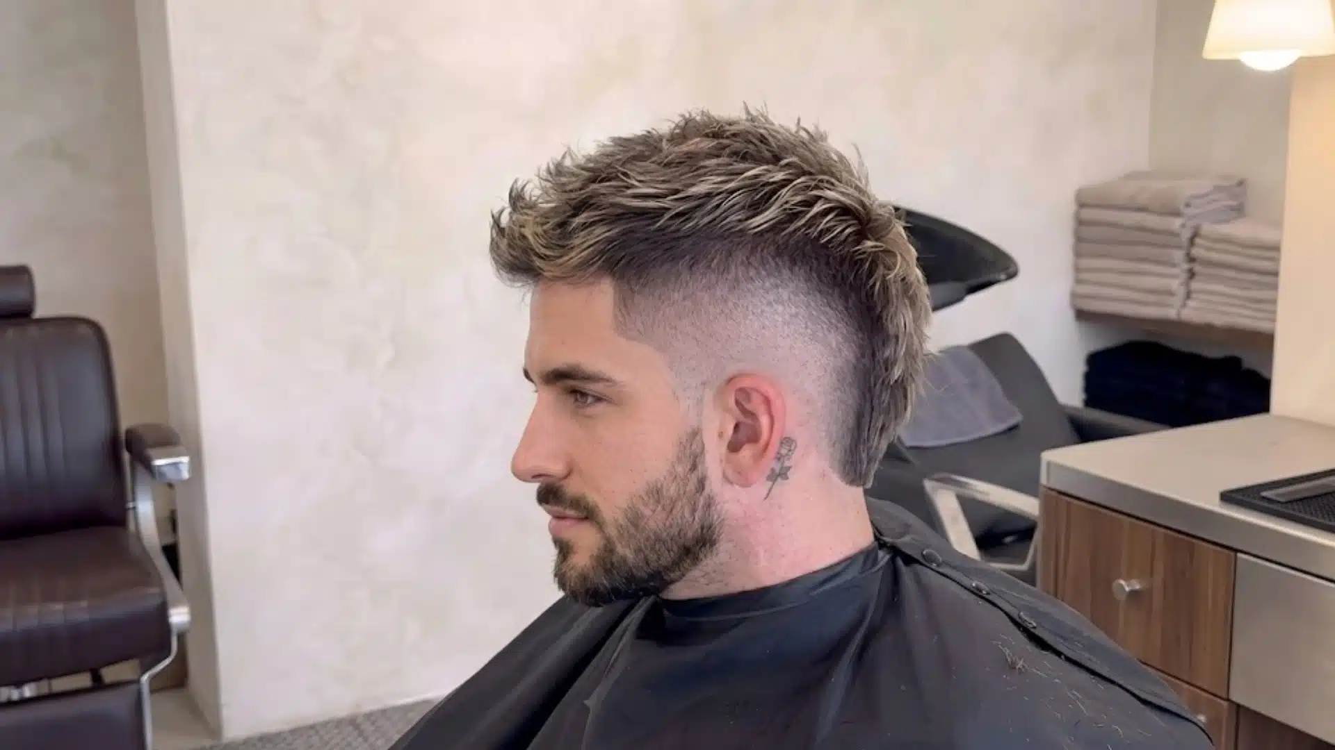 Profile of a man showing a textured burst fade mohawk haircut with blonde highlights in a barbershop