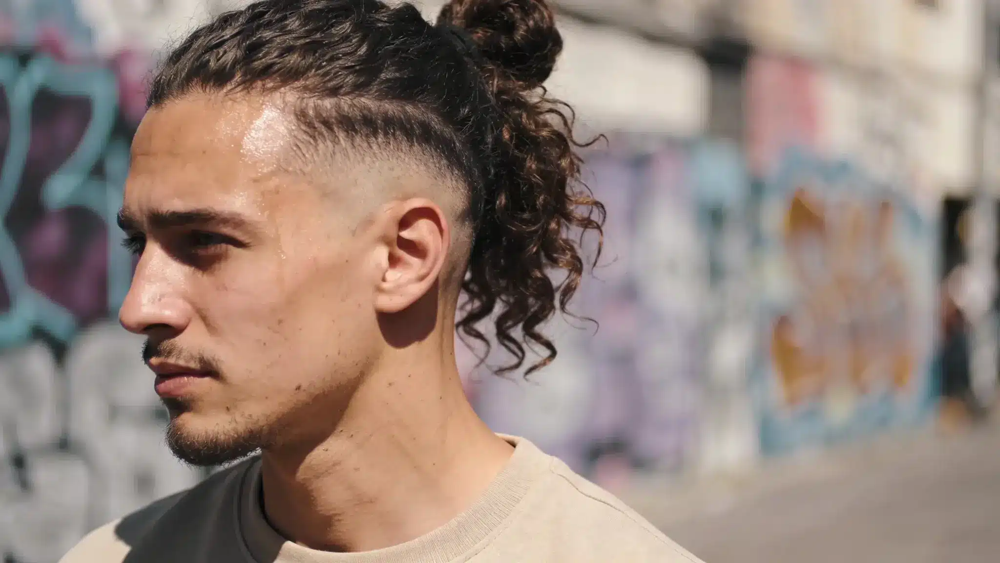 Profile of a man with a curly man bun and faded sides, standing outdoors against a blurred graffiti wall. webp