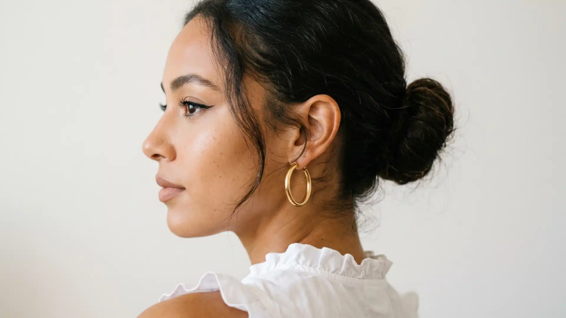 Profile of a woman with dark hair in a bun, wearing gold hoop earrings and a white ruffled top