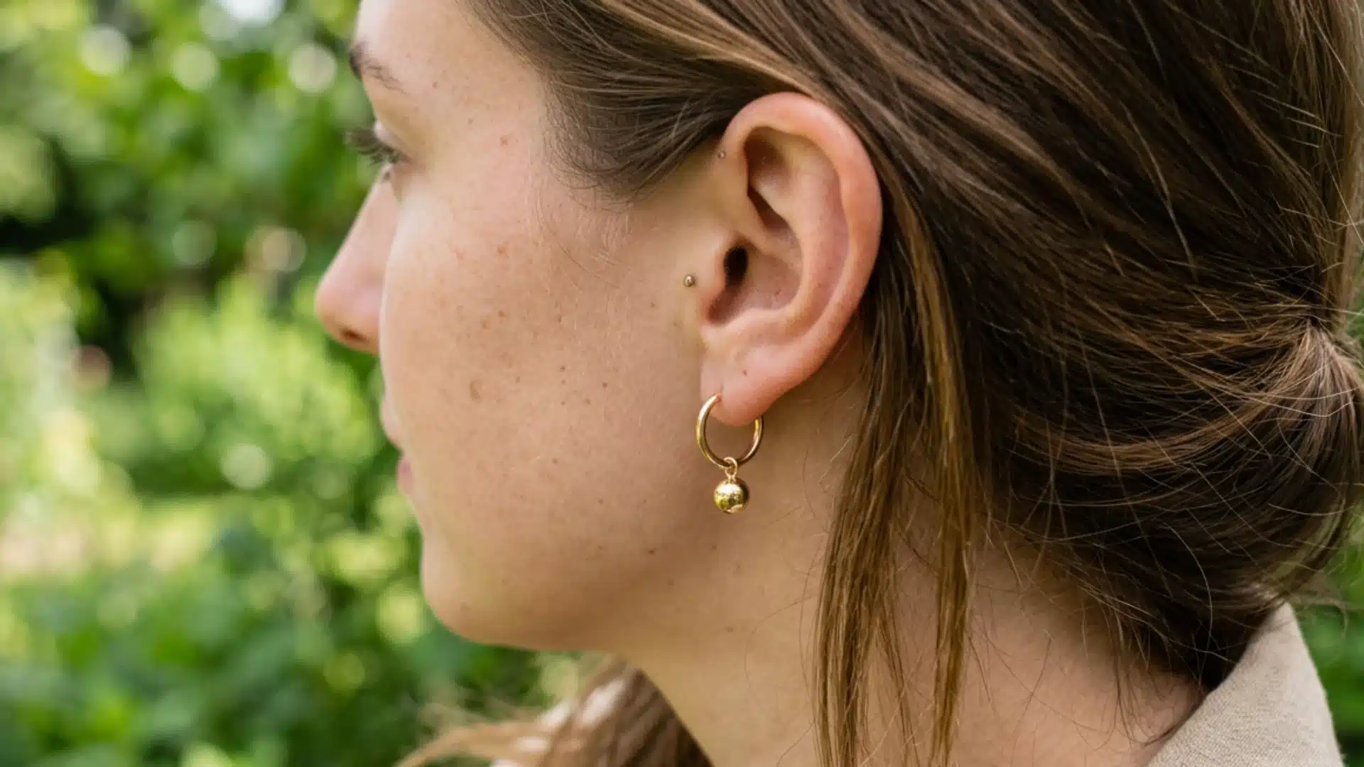 Profile of a woman with her hair tied back, wearing a small gold hoop earring with a dangling gold ball charm