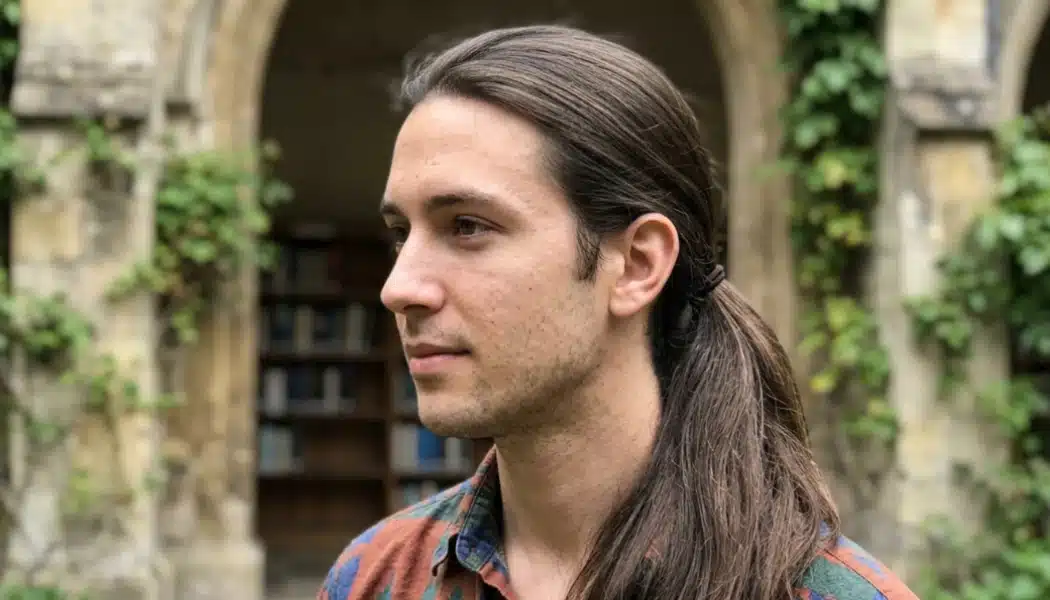 Side profile of a man with long straight hair in a low ponytail, standing in a historic stone courtyard with ivy-covered arches