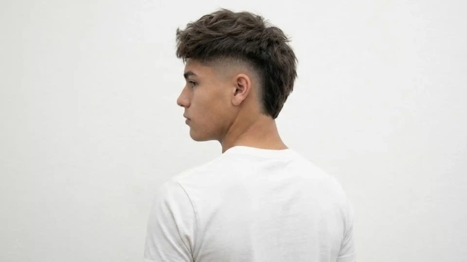 Side-profile of a young man with a skin fade mullet haircut against a plain white wall