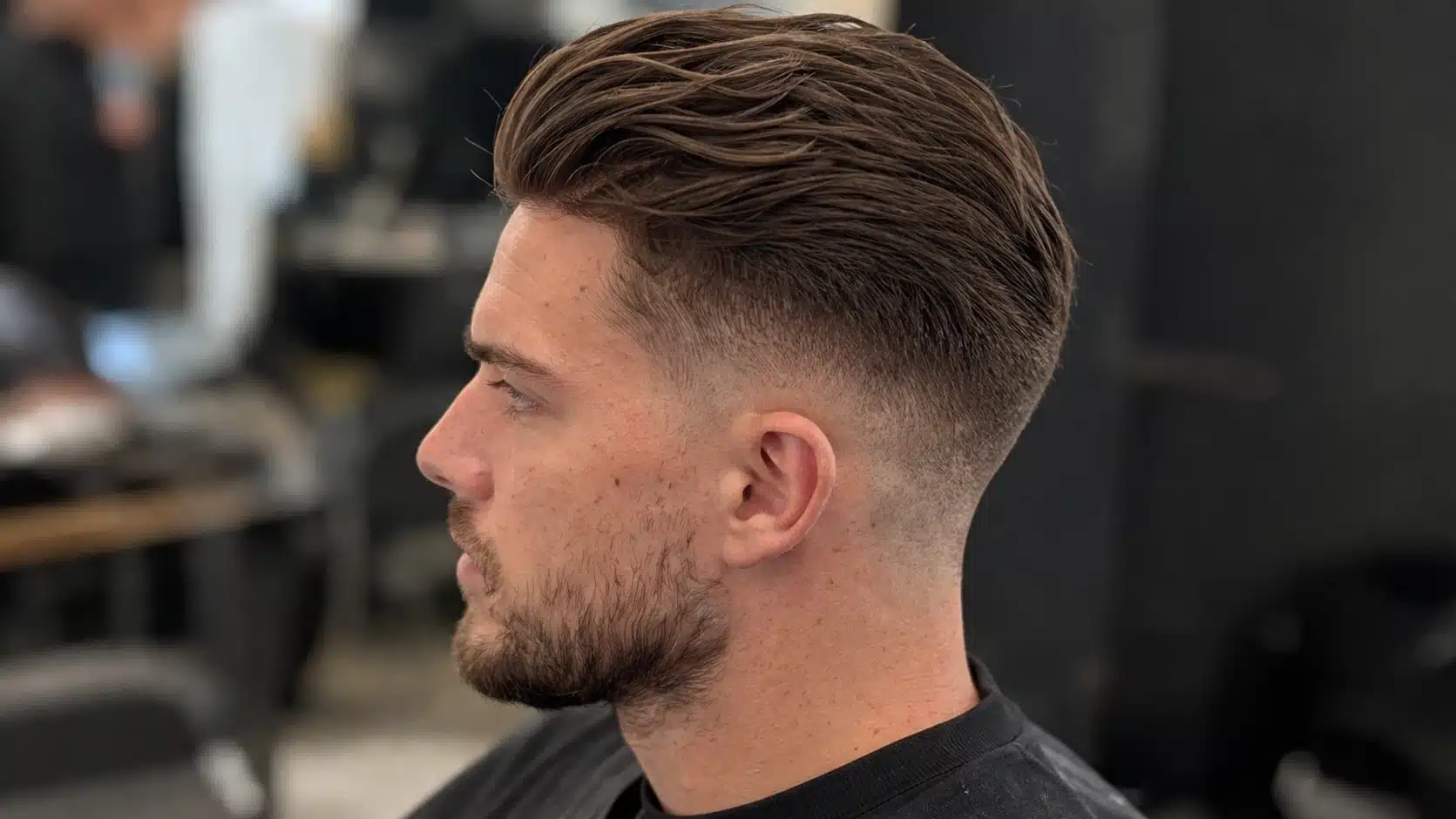 Side-profile portrait of a young man with a loose high fade slick back hairstyle and light stubble in a barbershop setting