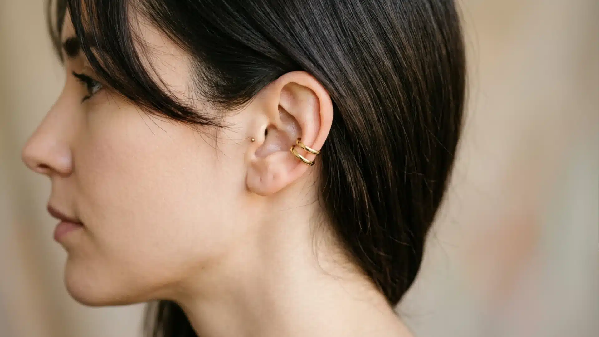 Side profile of a woman with dark hair and bangs, wearing a double-banded gold ear cuff and a tiny tragus stud