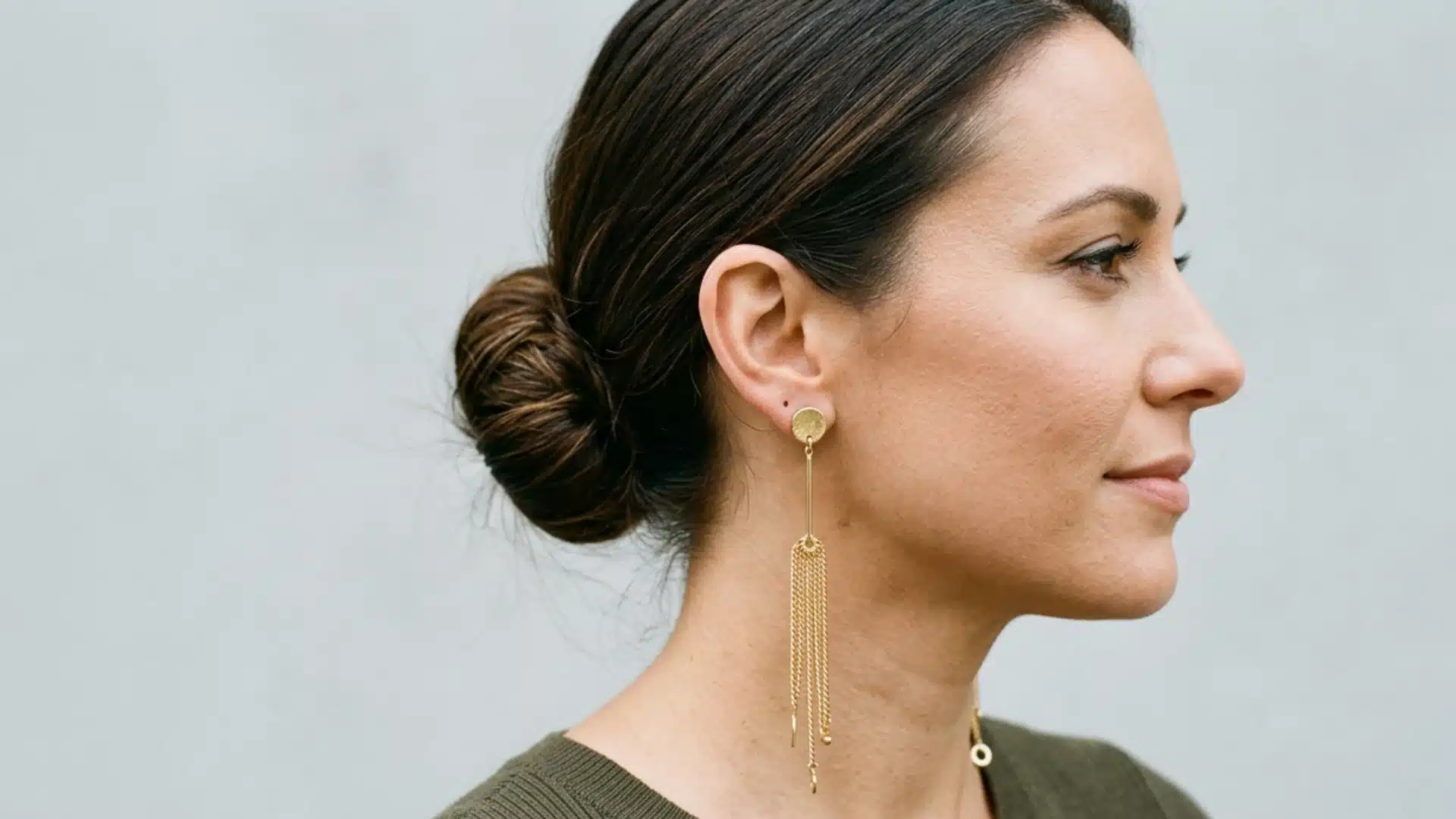 Side profile of a woman with dark hair in a bun, wearing long gold drop earrings with a tassel of fine chains.wep