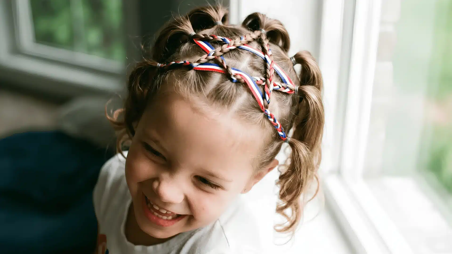 Smiling girl with colorful braided hair and ribbons by a window