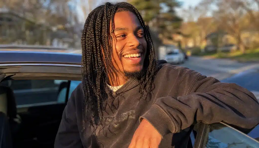 Smiling man with long box braids leaning against a car at golden hour