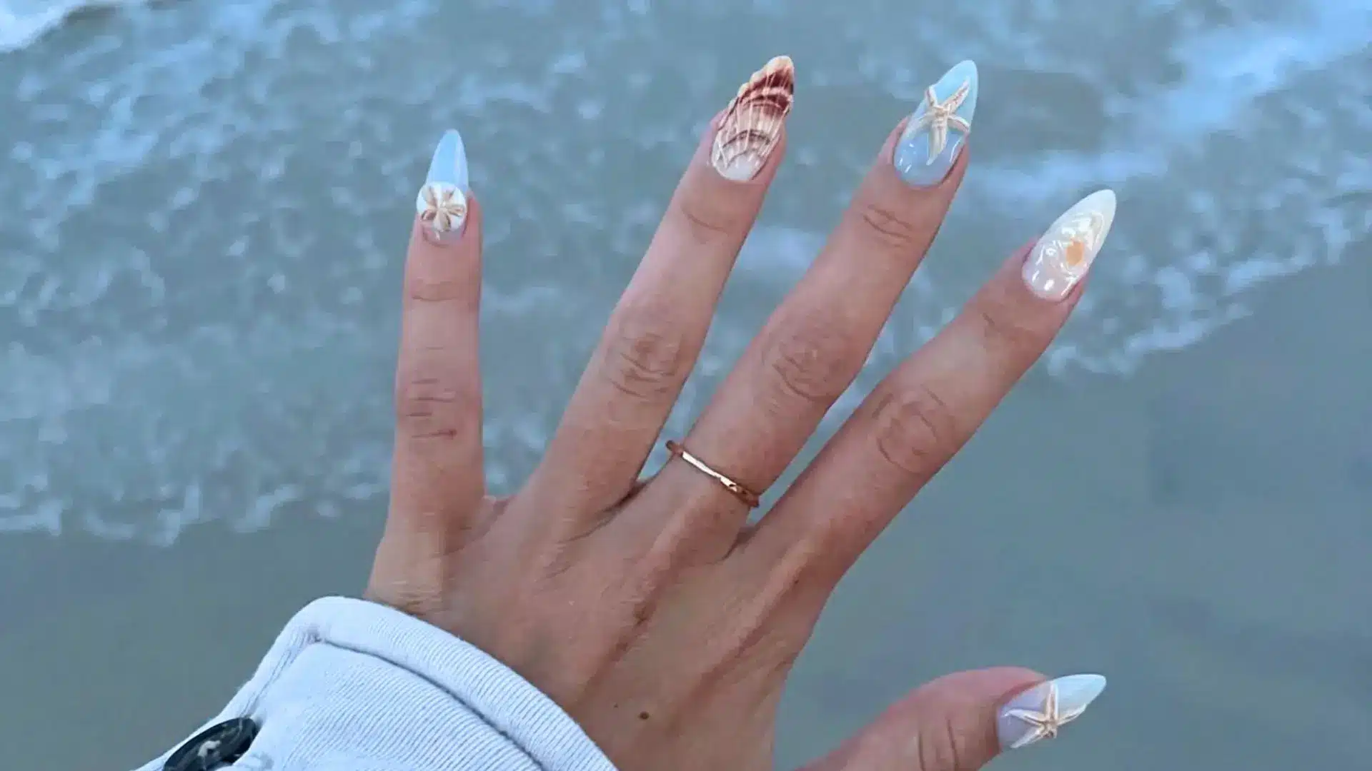 Starfish and seashell nail art on almond nails in soft blue and nude tones with ocean water background