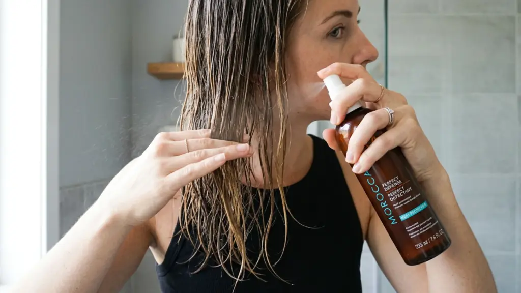 Woman applying Moroccanoil heat protectant spray to damp hair before styling in a bathroom