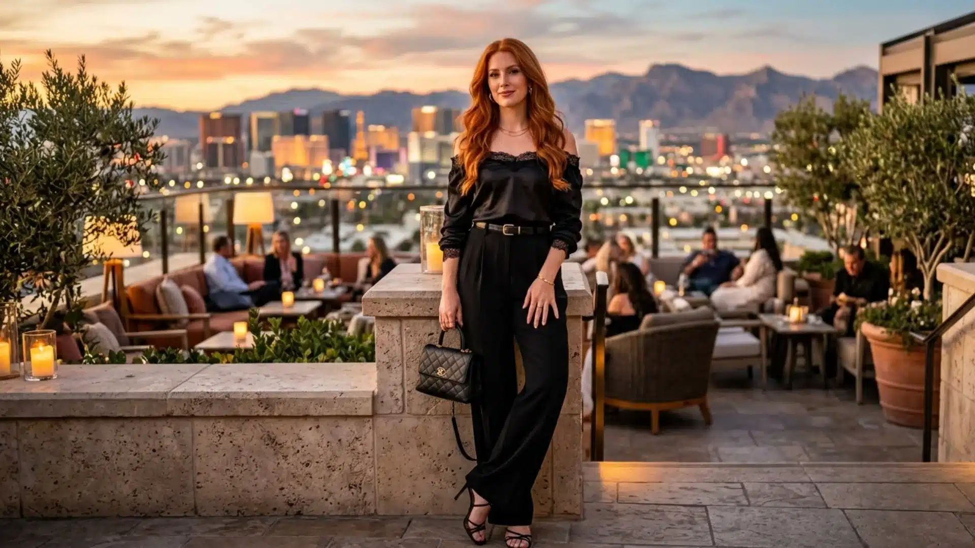 Woman in black evening outfit holding a handbag on a rooftop terrace with city lights and mountains behind her