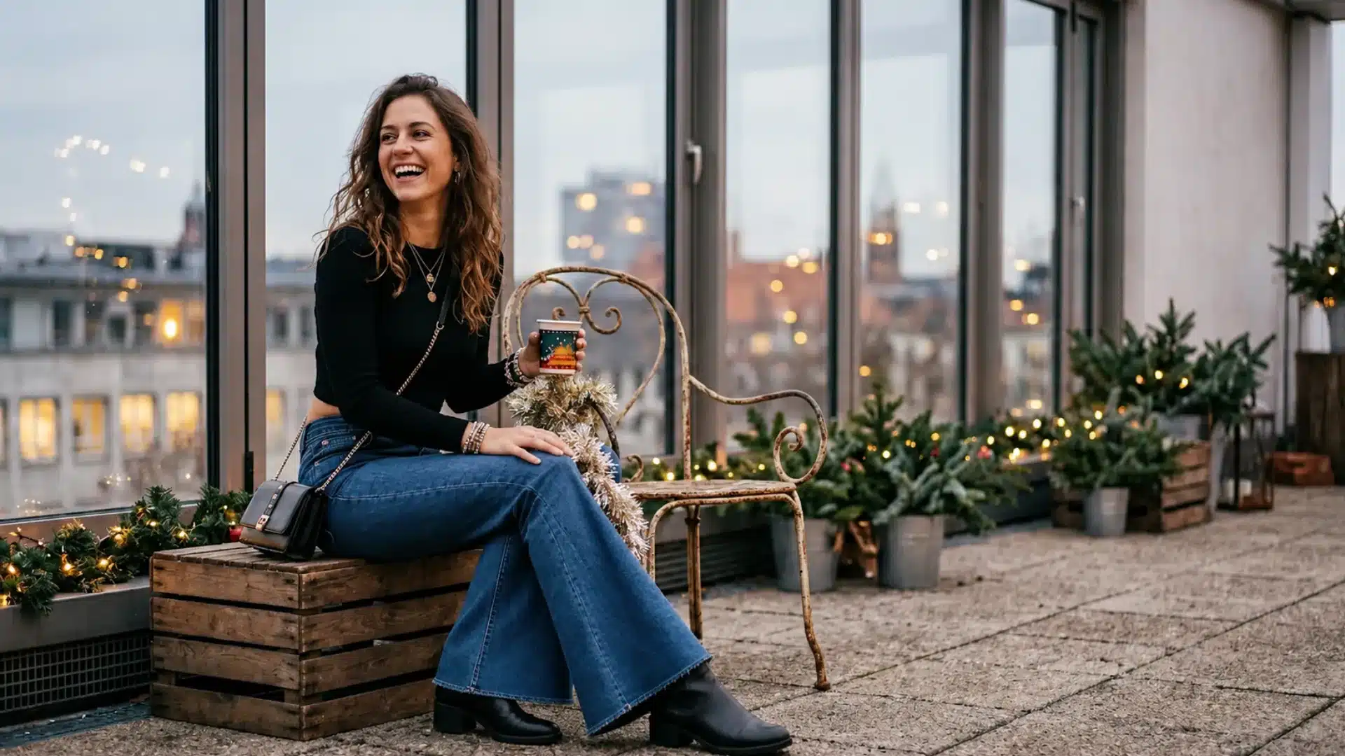 Woman in black top, blue flare jeans, and black boots sitting on a rooftop terrace at dusk