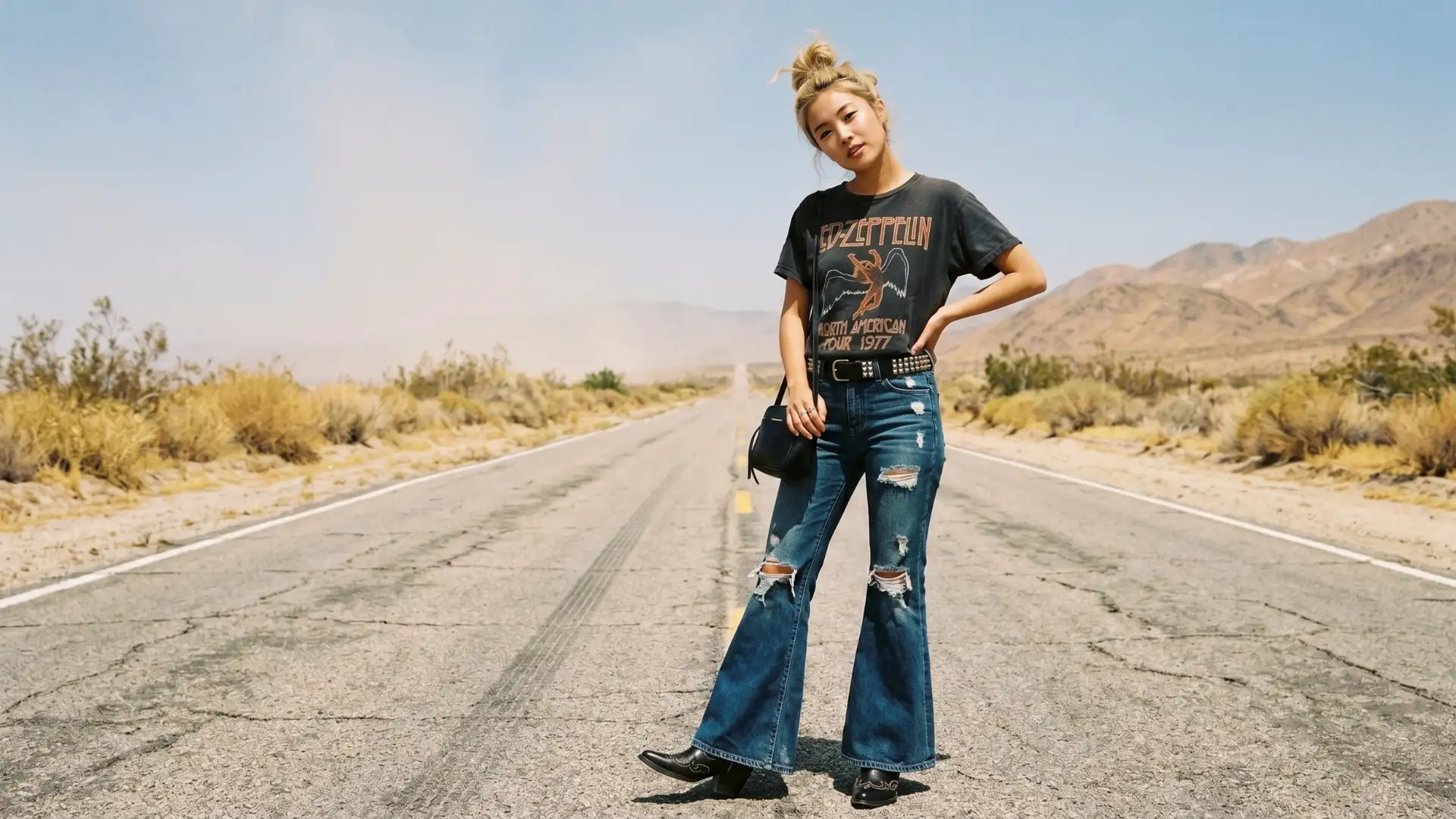 Woman in graphic tee, flare jeans, and western-inspired boots posing on a desert road landscape