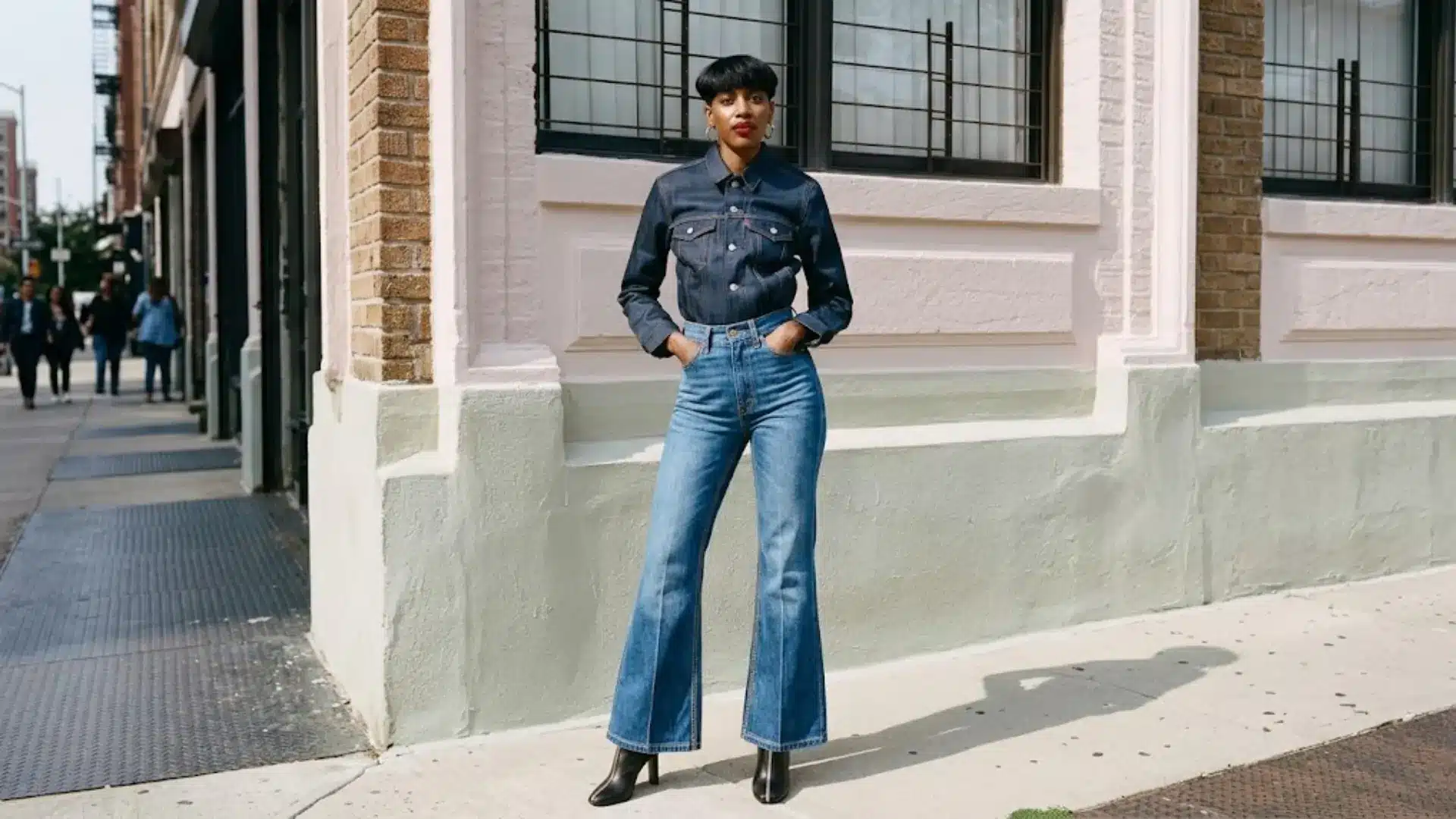 Woman in wide flare jeans and denim shirt posing on a sunny urban street in black ankle boots