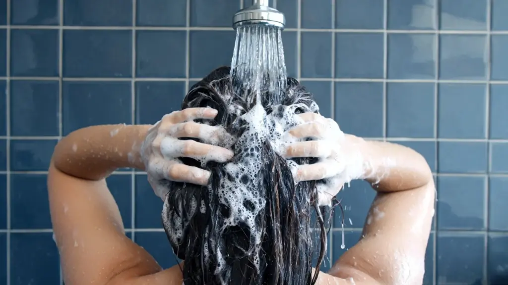 Woman washing her hair under a showerhead while massaging shampoo into her scalp with both hands in a tiled bathroom