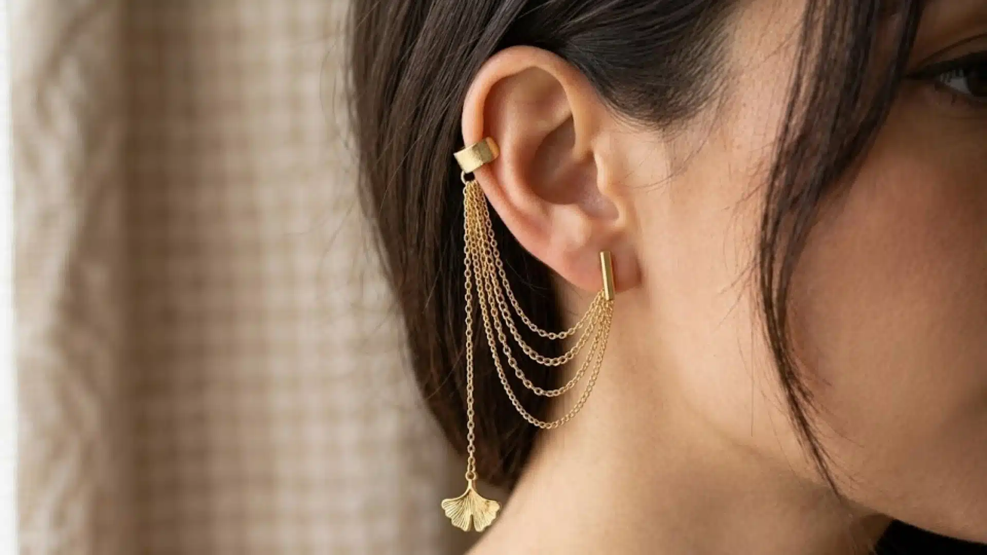 Woman wearing a gold chain ear cuff connected to a stud earring with layered draped chains and charm