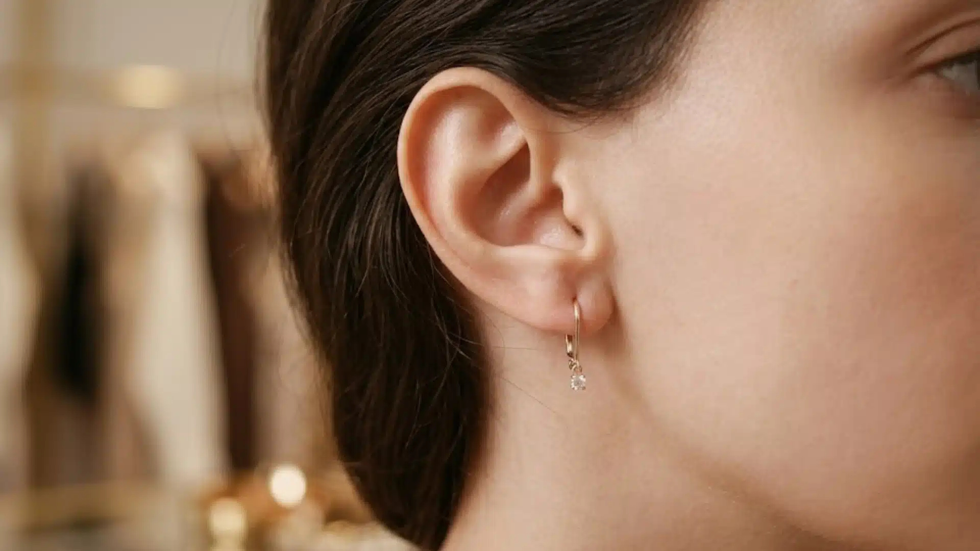 Woman wearing a small gold leverback hoop earring with a tiny crystal drop in a soft indoor setting