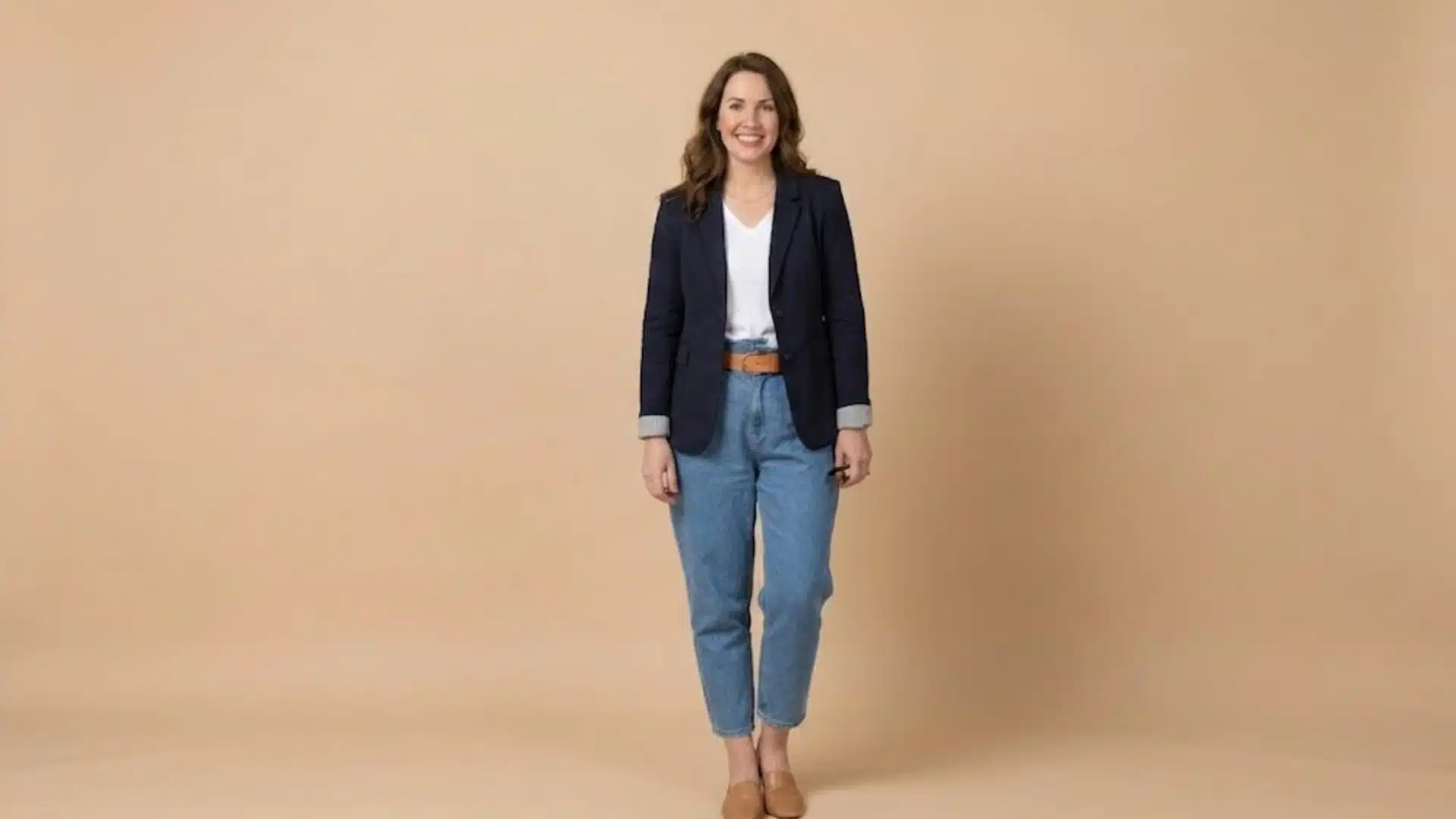 Woman wearing paperbag waist jeans with navy blazer, white top, and tan flats against beige background