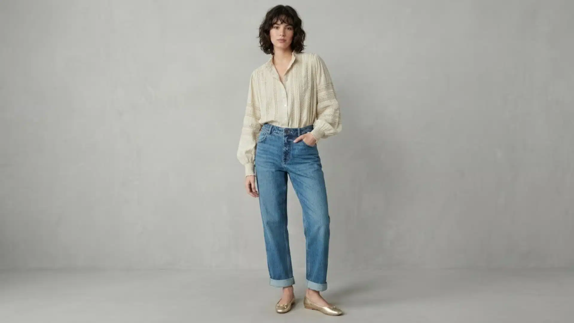 Woman wearing relaxed boyfriend jeans with tucked beige blouse and gold flats in minimal studio setting