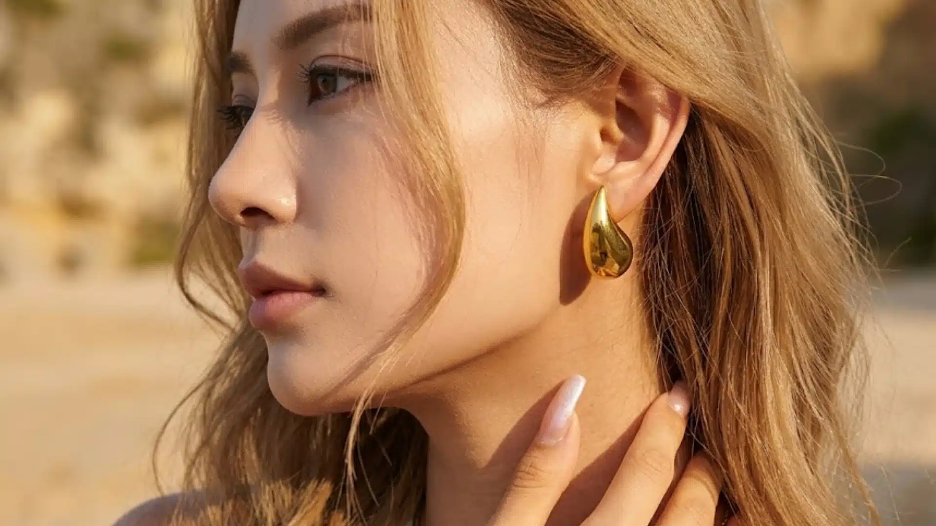 Woman wearing shiny gold teardrop earrings in warm sunlight with soft waves and a beach backdrop