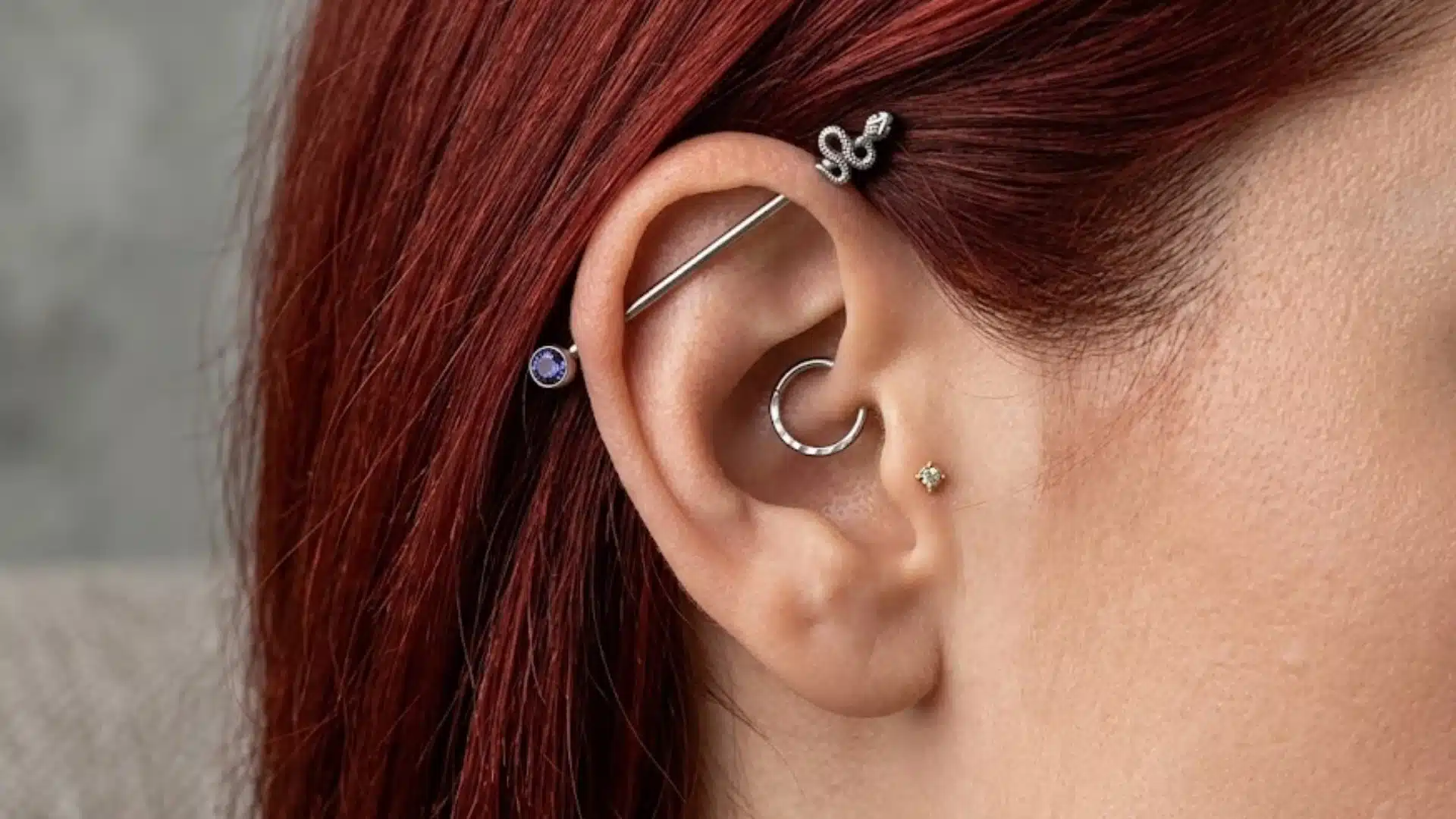 Woman wearing silver barbell earrings across the upper ear with small studs and red hair against a gray backdrop