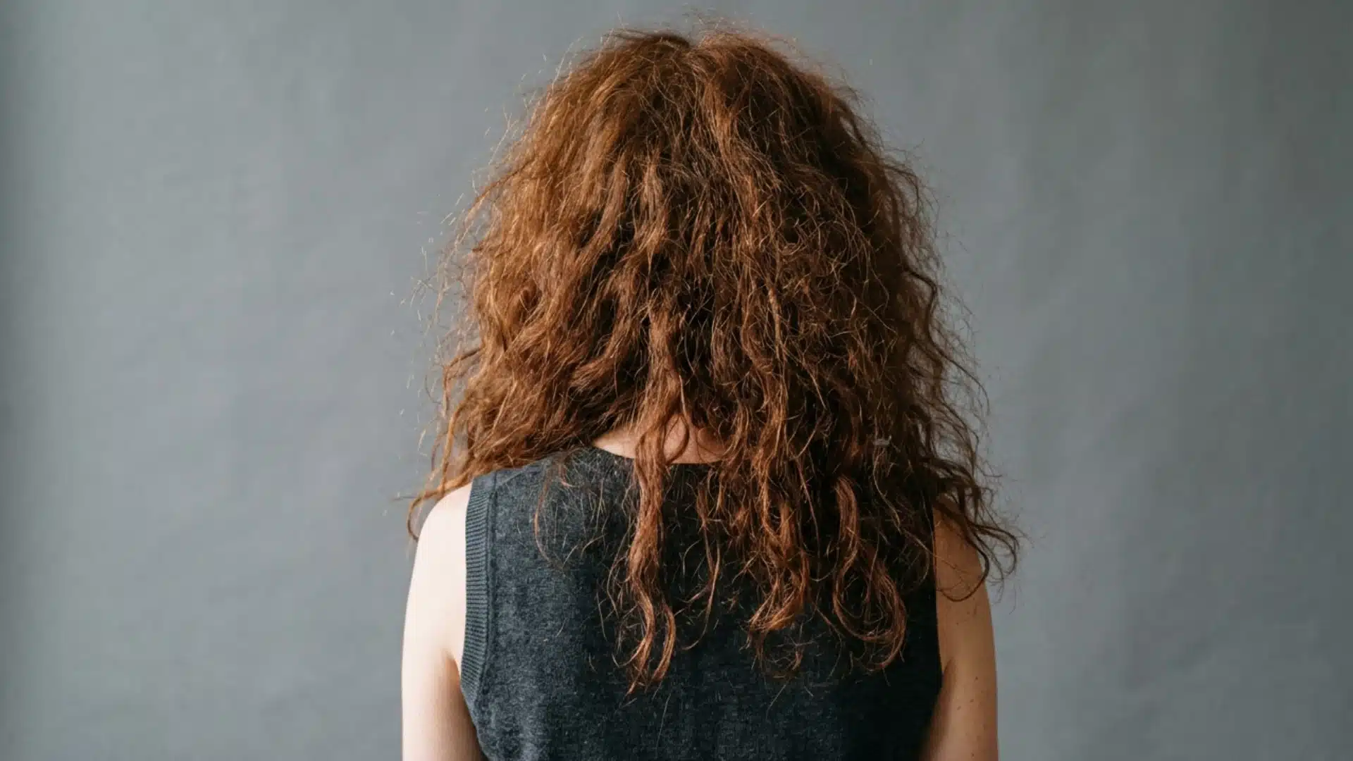 Woman with frizzy wavy auburn hair shown from the back against a grey background, highlighting dry texture and flyaways