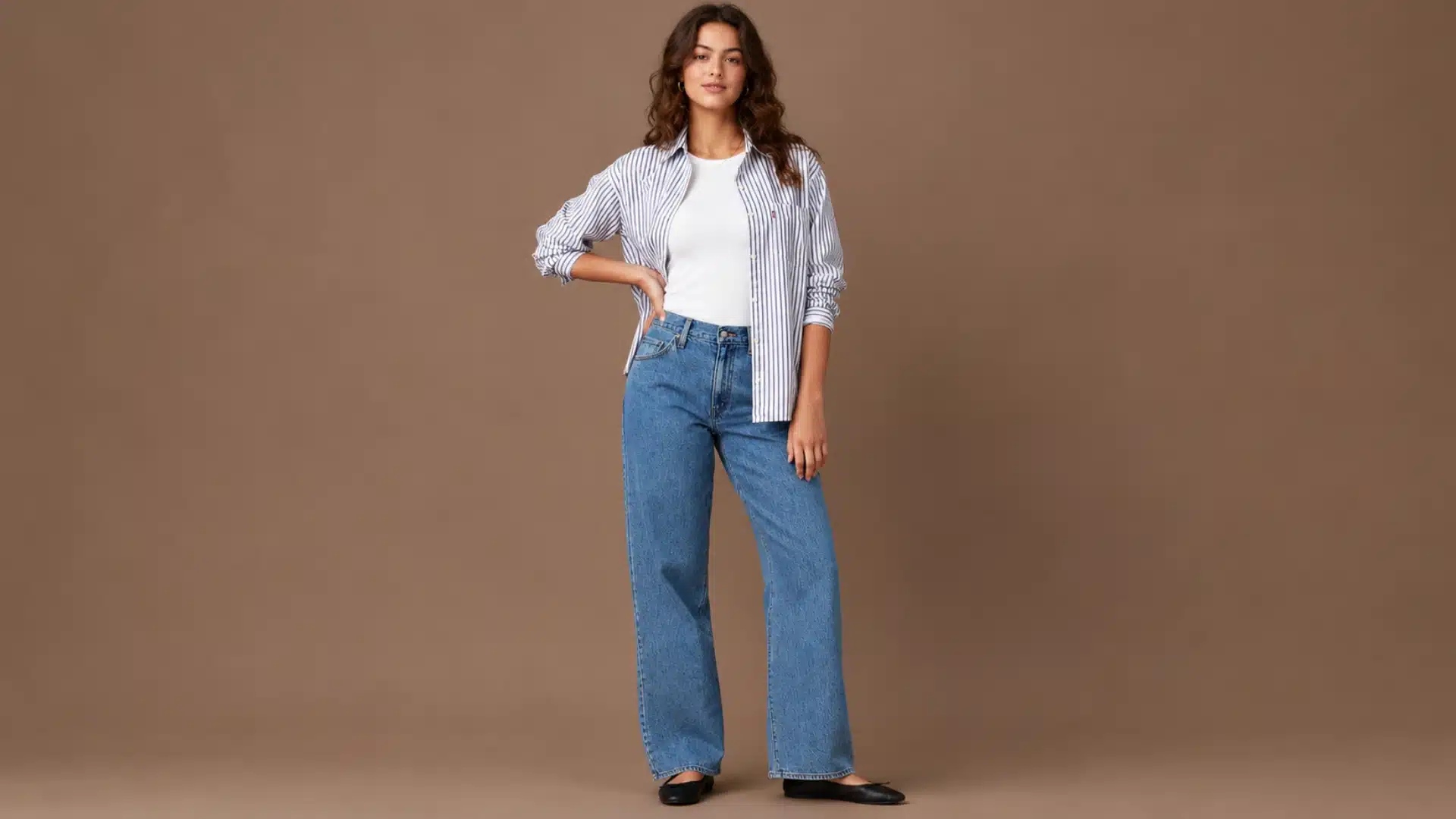 Woman in a striped button-down shirt, white top, and blue wide-leg jeans against a brown backdrop