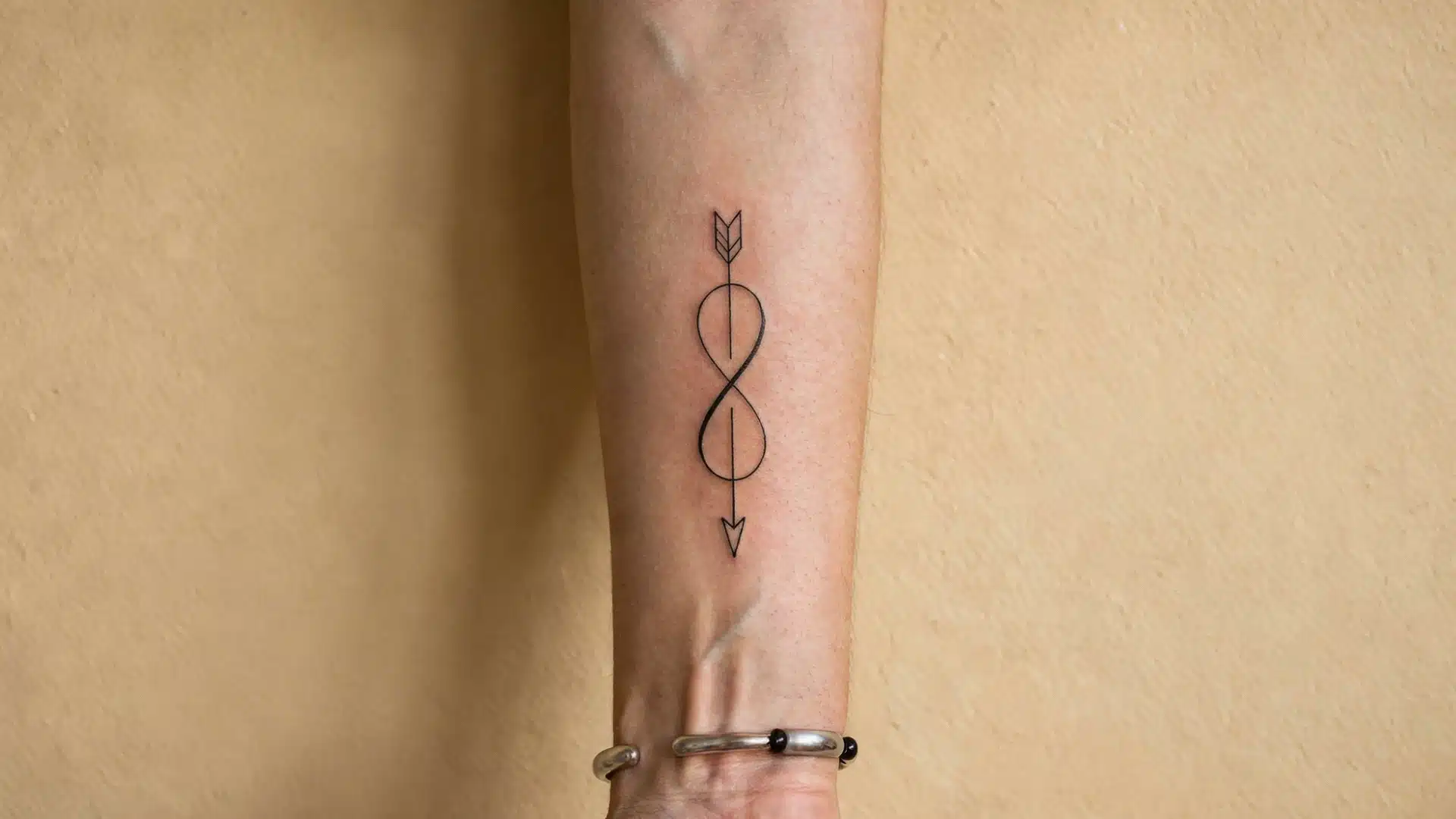 A fine-line tattoo of an arrow woven through an infinity symbol on a person's inner forearm, pointing down