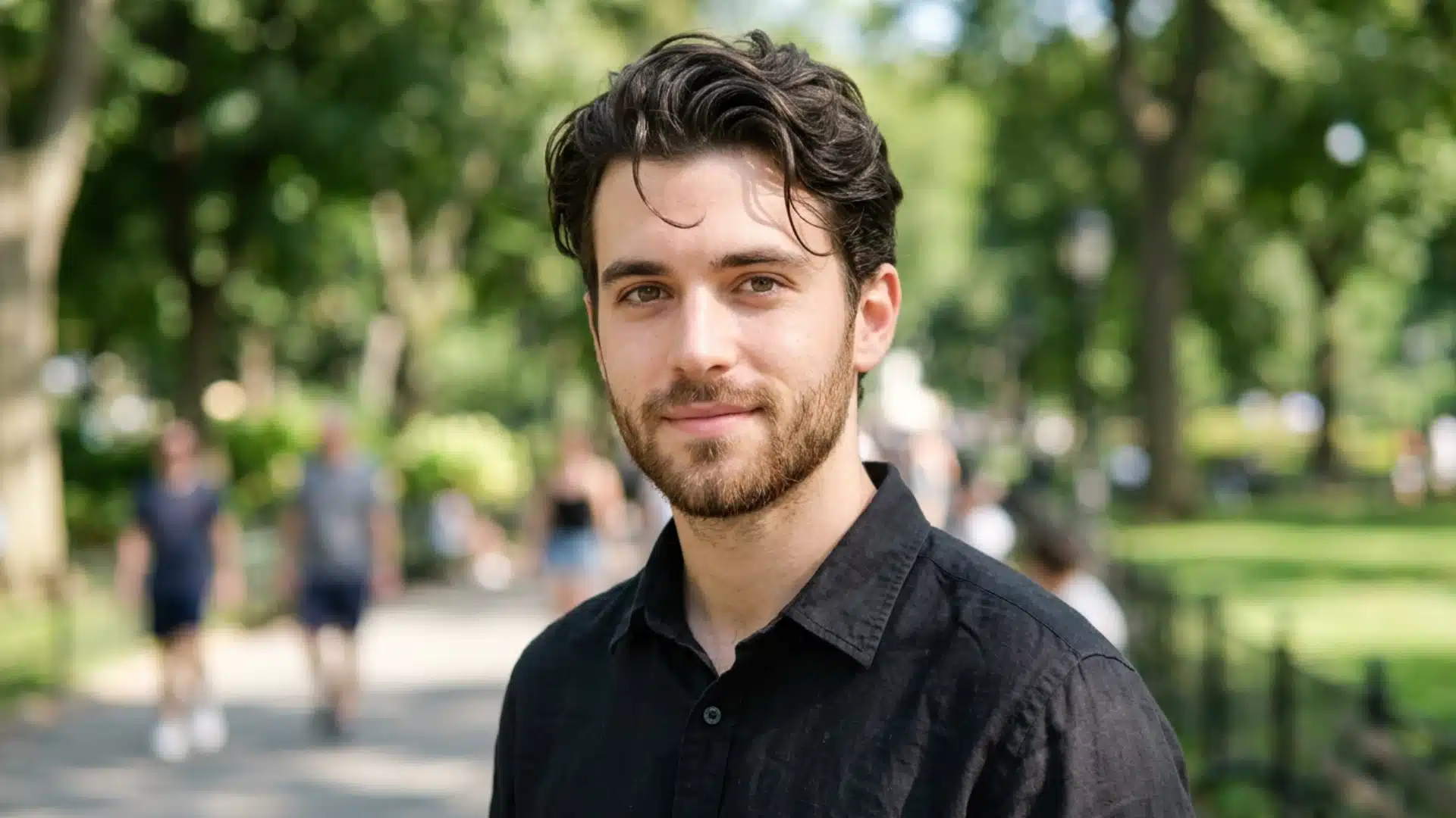 A portrait of a man with dark, wavy hair and a trimmed beard smiling slightly in a sunny, blurred park setting