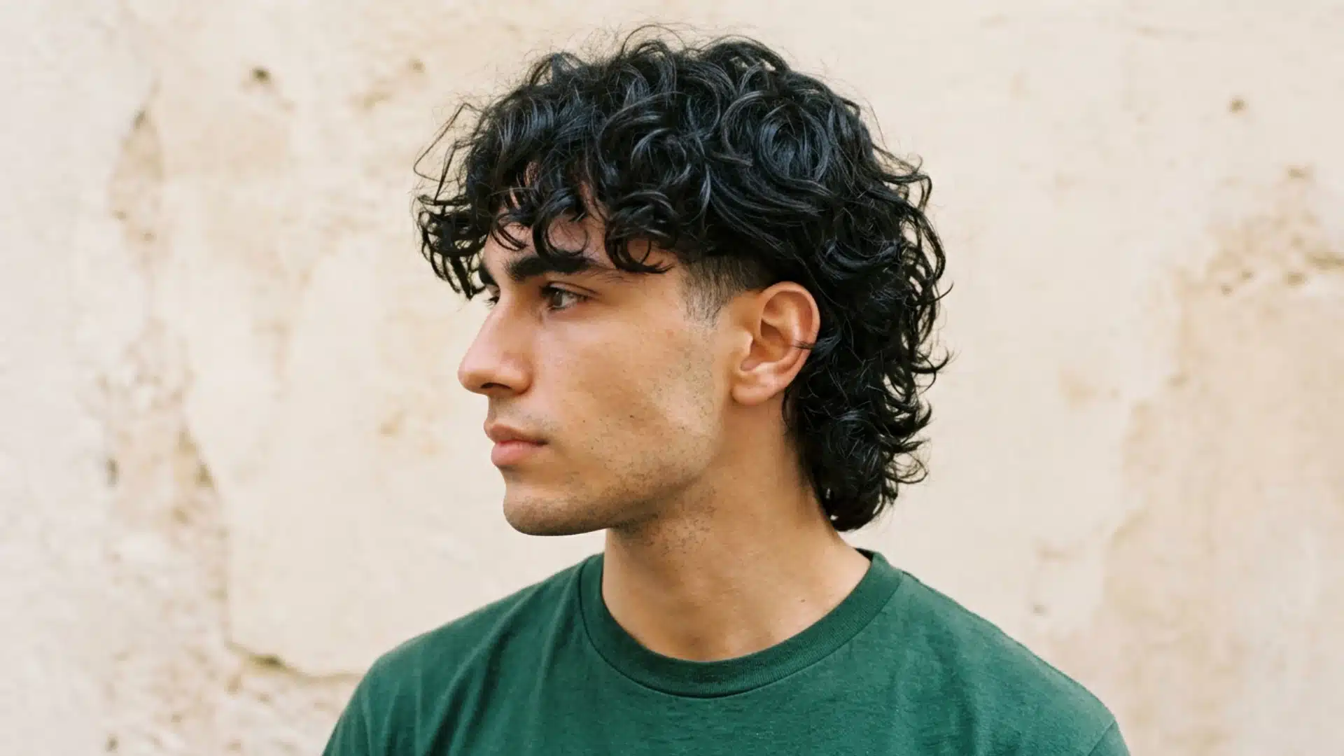 A profile photo of a young man with dark, curly hair and a green t-shirt against a textured wall