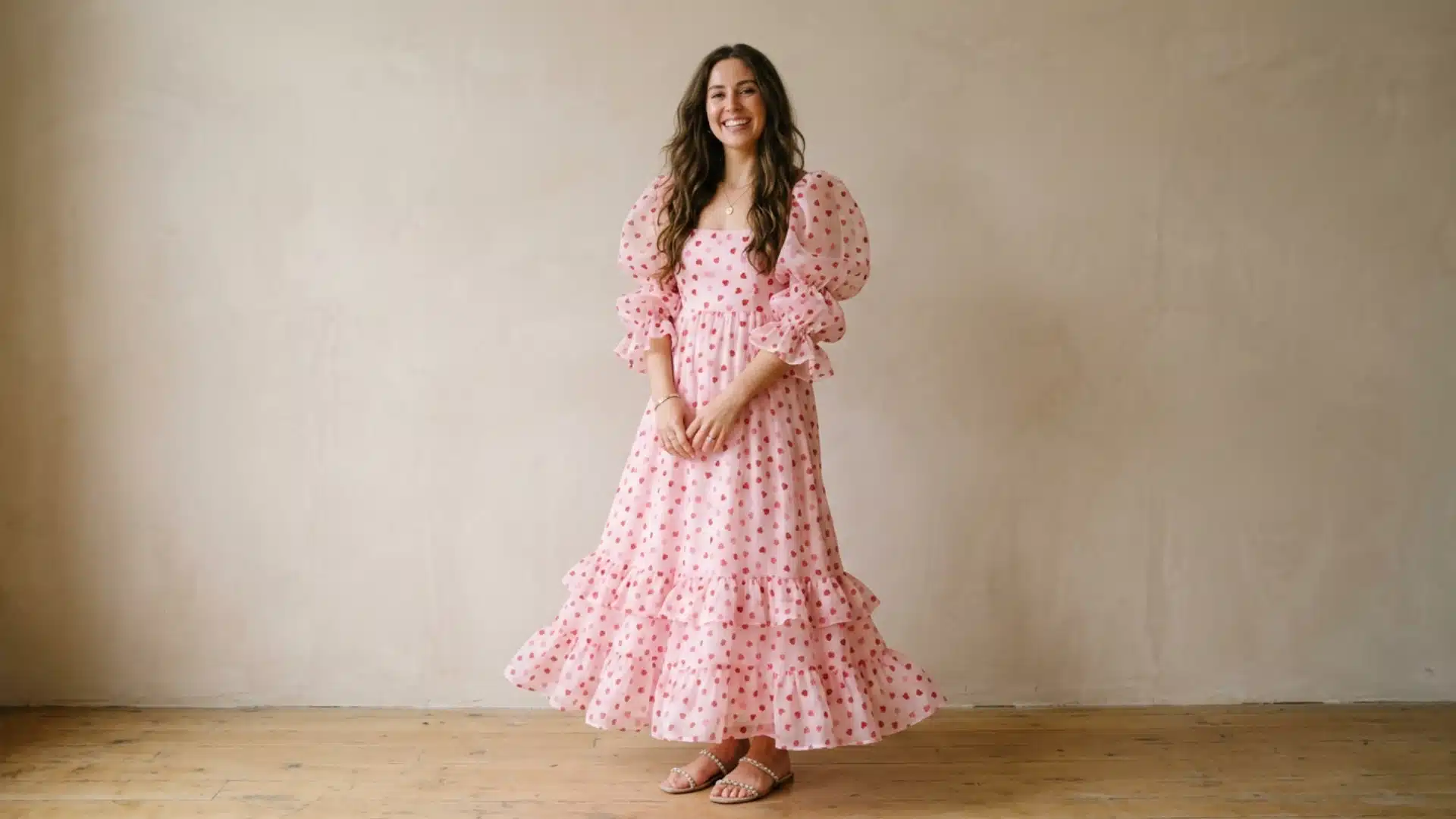 A smiling woman poses in a light pink, tiered maxi dress patterned with small red hearts and featuring puff sleeves