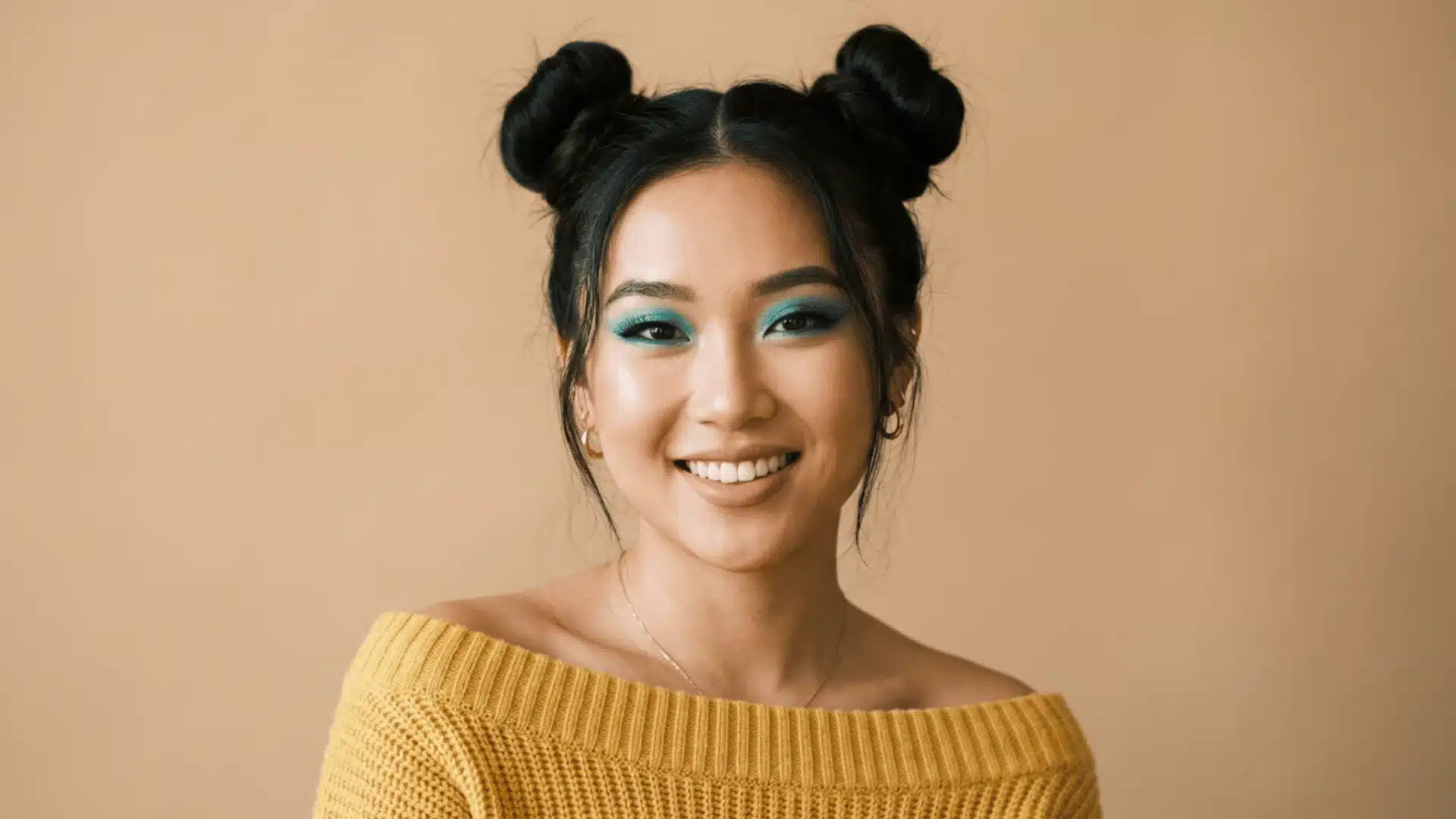 A smiling woman wearing a yellow top has her hair styled into two space buns, featuring vibrant teal eyeshadow