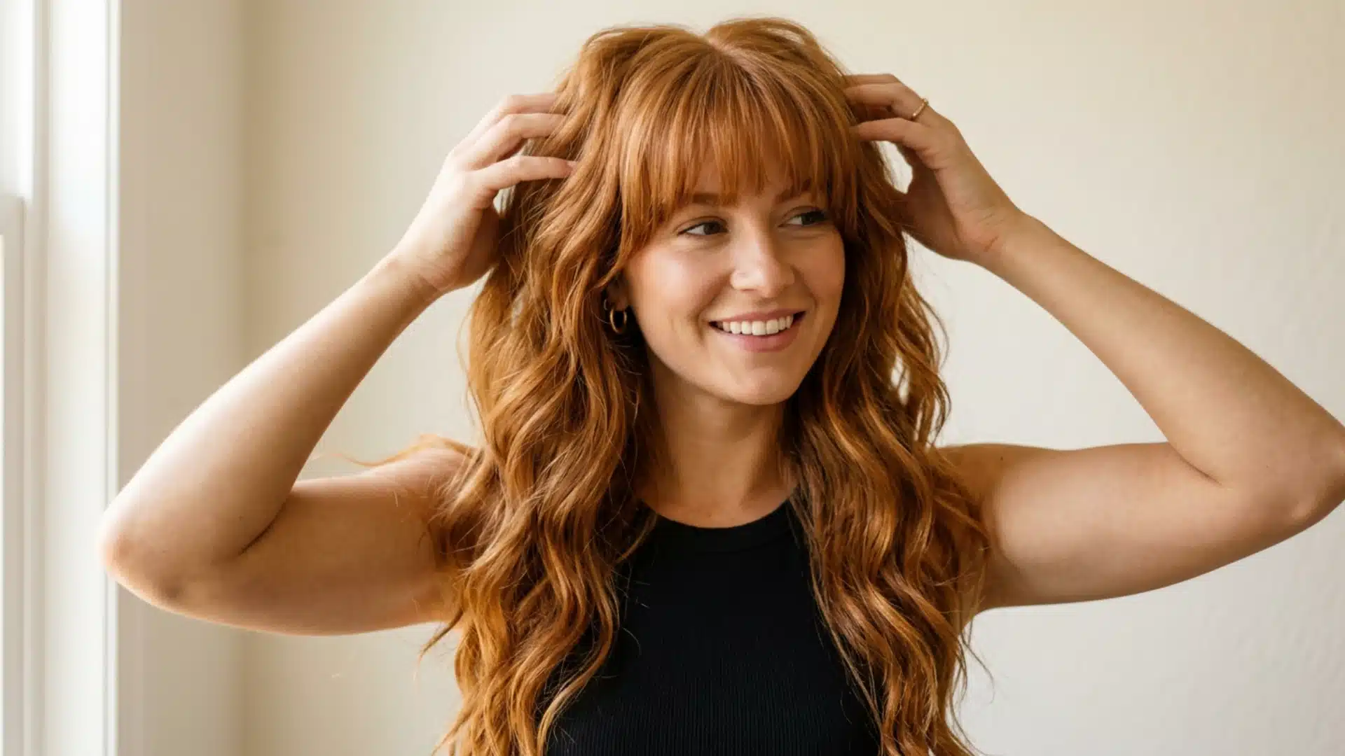 A smiling young woman with long, wavy copper red hair running her hands through her hair