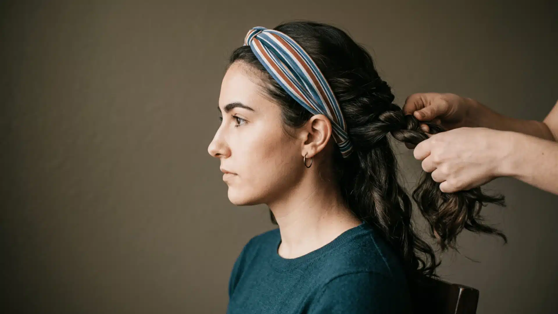 A woman with a striped headband has her long, dark wavy hair being braided from behind by someone's hands