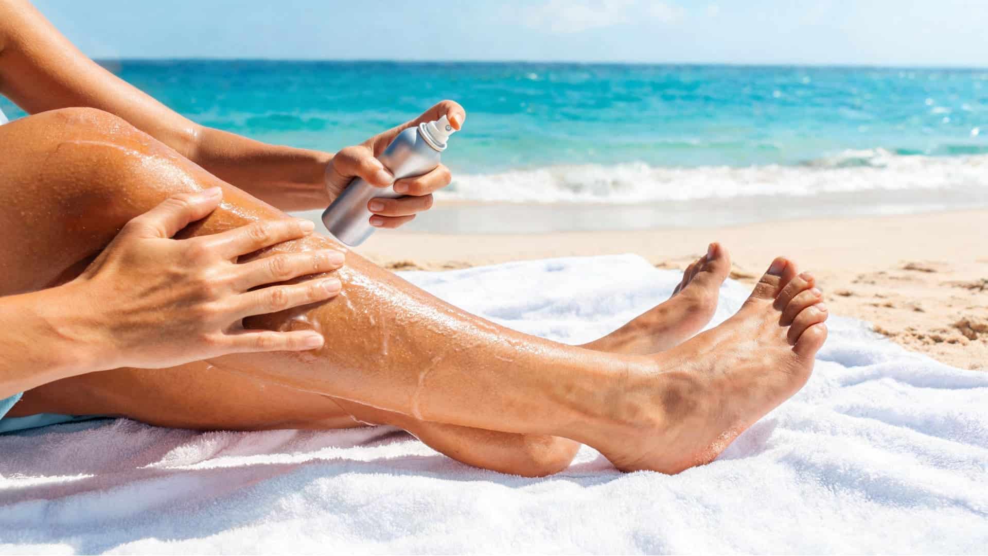 A person applying tanning oil from a spray bottle onto their legs while relaxing on a beach towel by the ocean