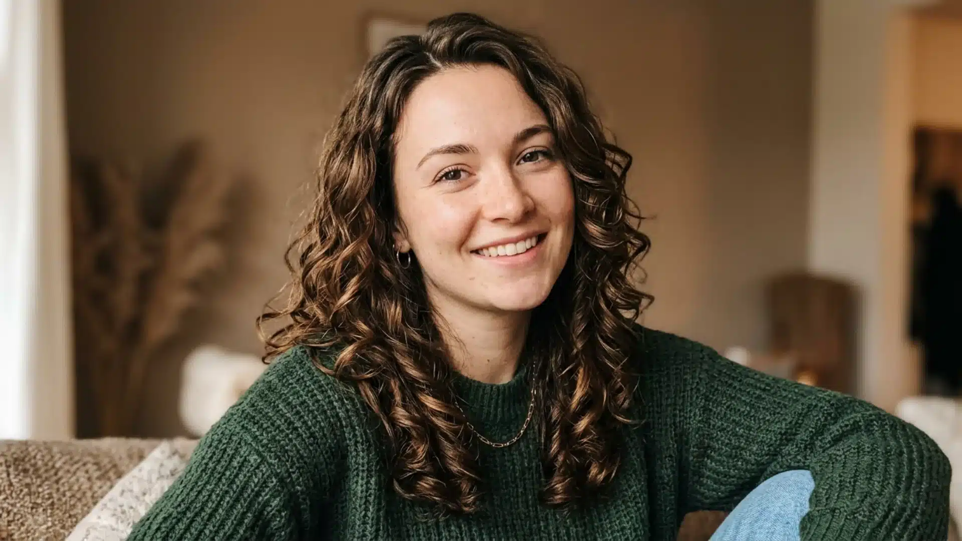 A smiling woman with curly brown hair wears a green knit sweater in