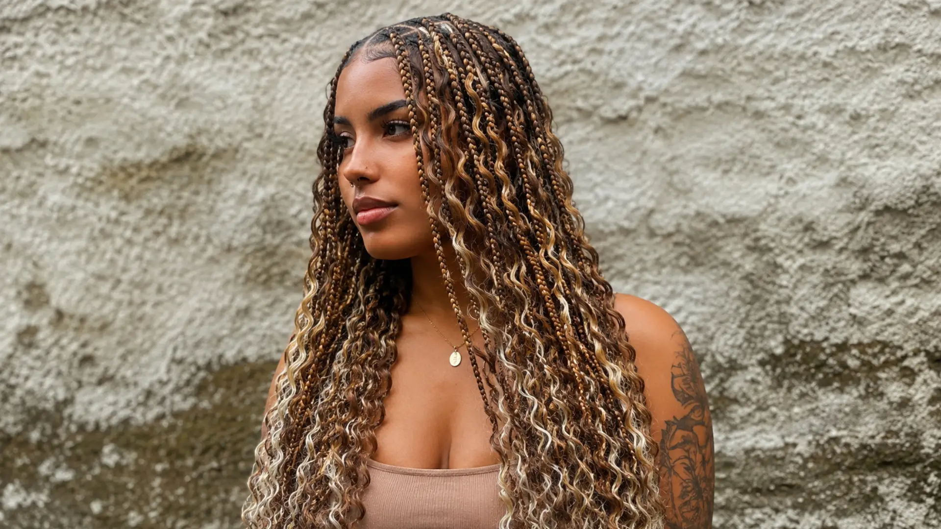 A woman with long, middle-parted goddess box braids in shades of brown and blonde, showing loose curly strands