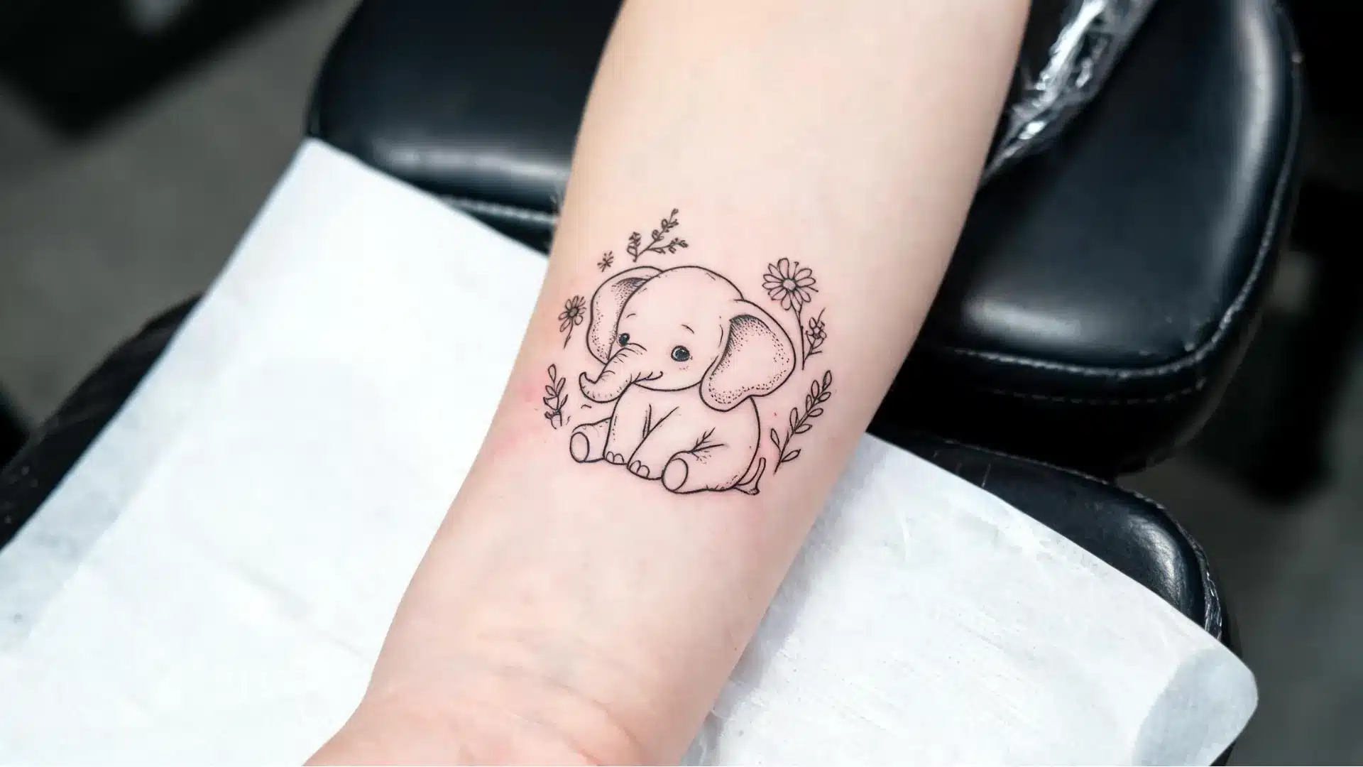 Baby elephant fine-line tattoo with small floral details on the inner forearm in a tattoo studio