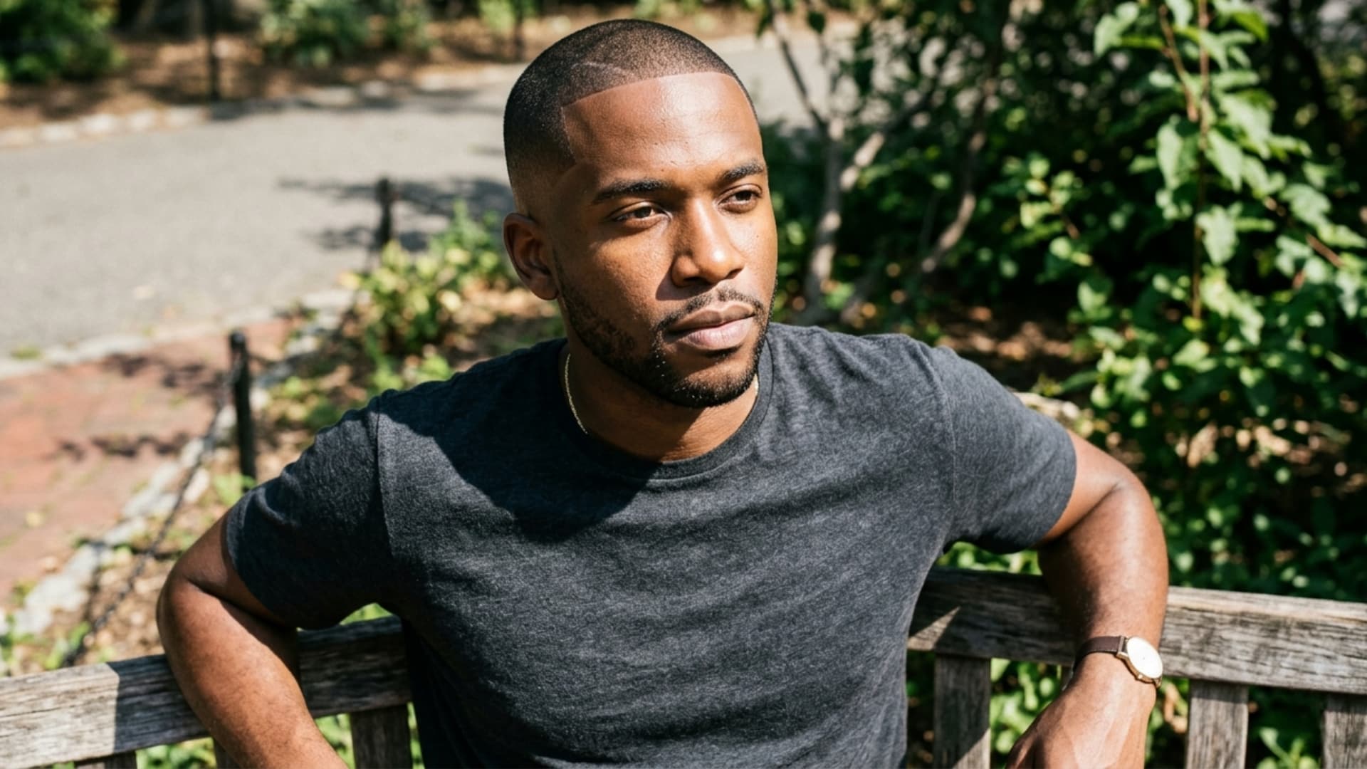 Black man with a sharp buzz hairstyle and short beard sitting on a wooden bench outdoors