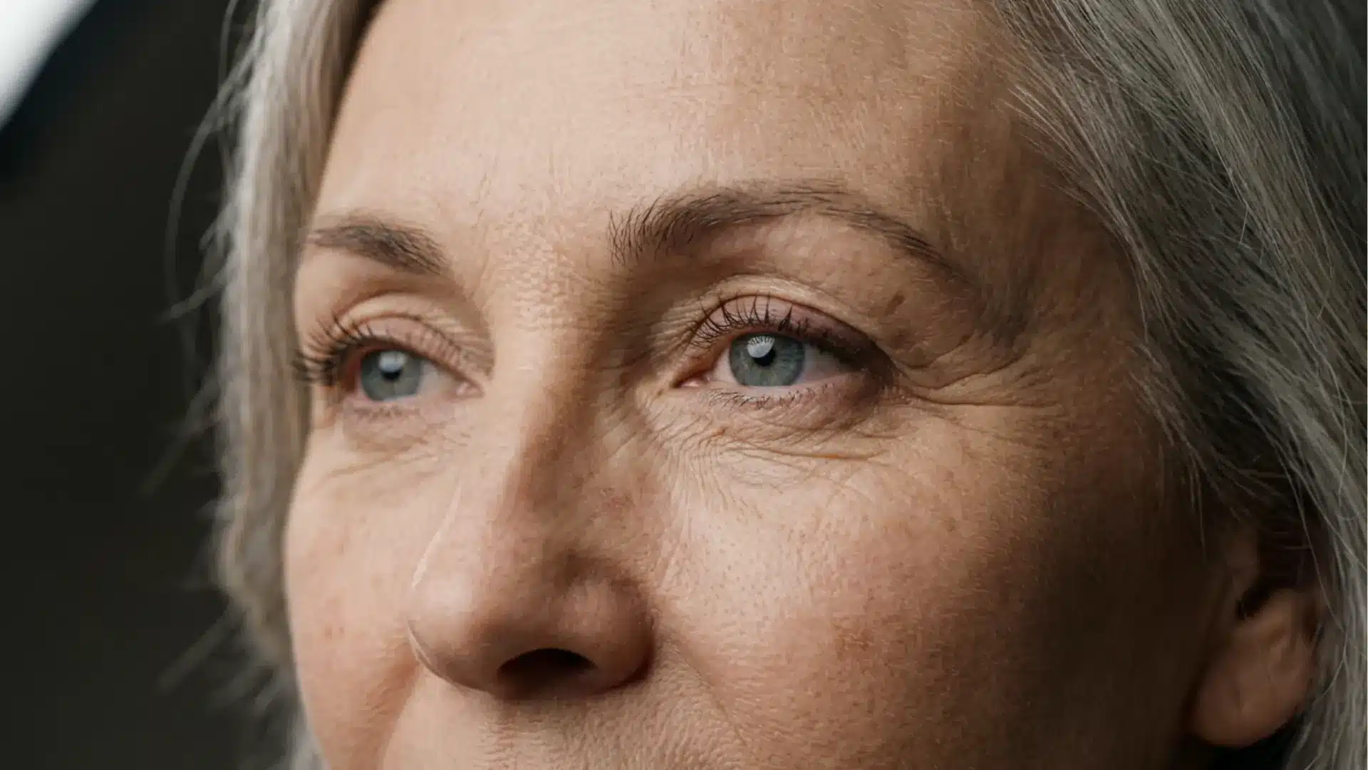 Close-up of an older woman's face showing fine lines around her blue eyes and graying hair, looking off-camera