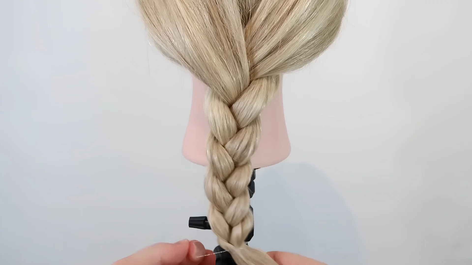 Finished blonde three-strand braid being secured at the end with a small clear elastic to hold the hairstyle in place.