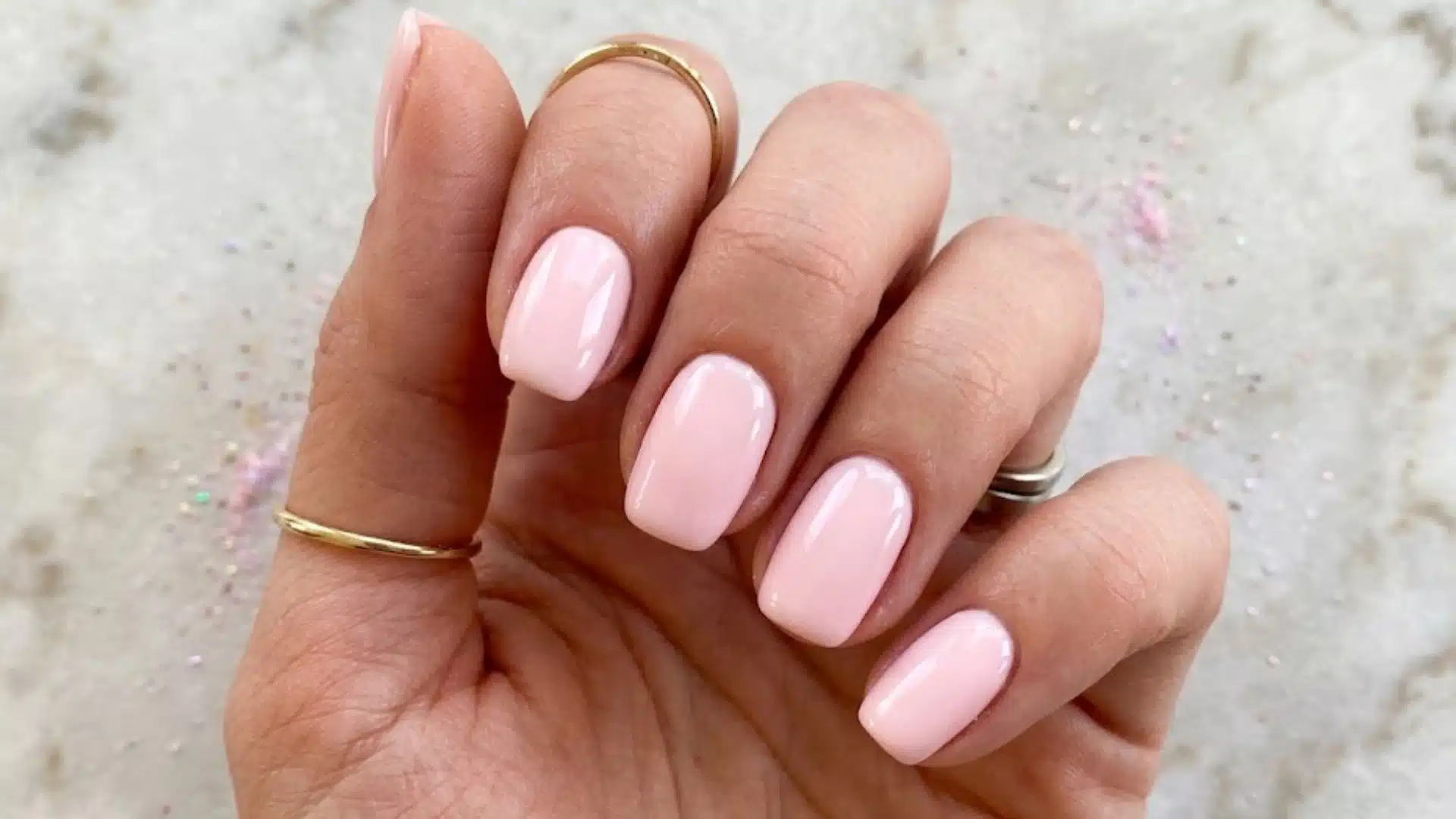Glossy light pink square nails on hand with gold rings against a soft marble background with subtle sparkle