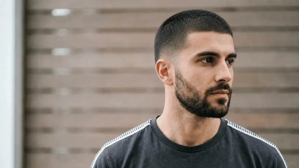 Guy with a defined buzz cut, faded sides, and trimmed beard looking sideways against a blurred wooden background