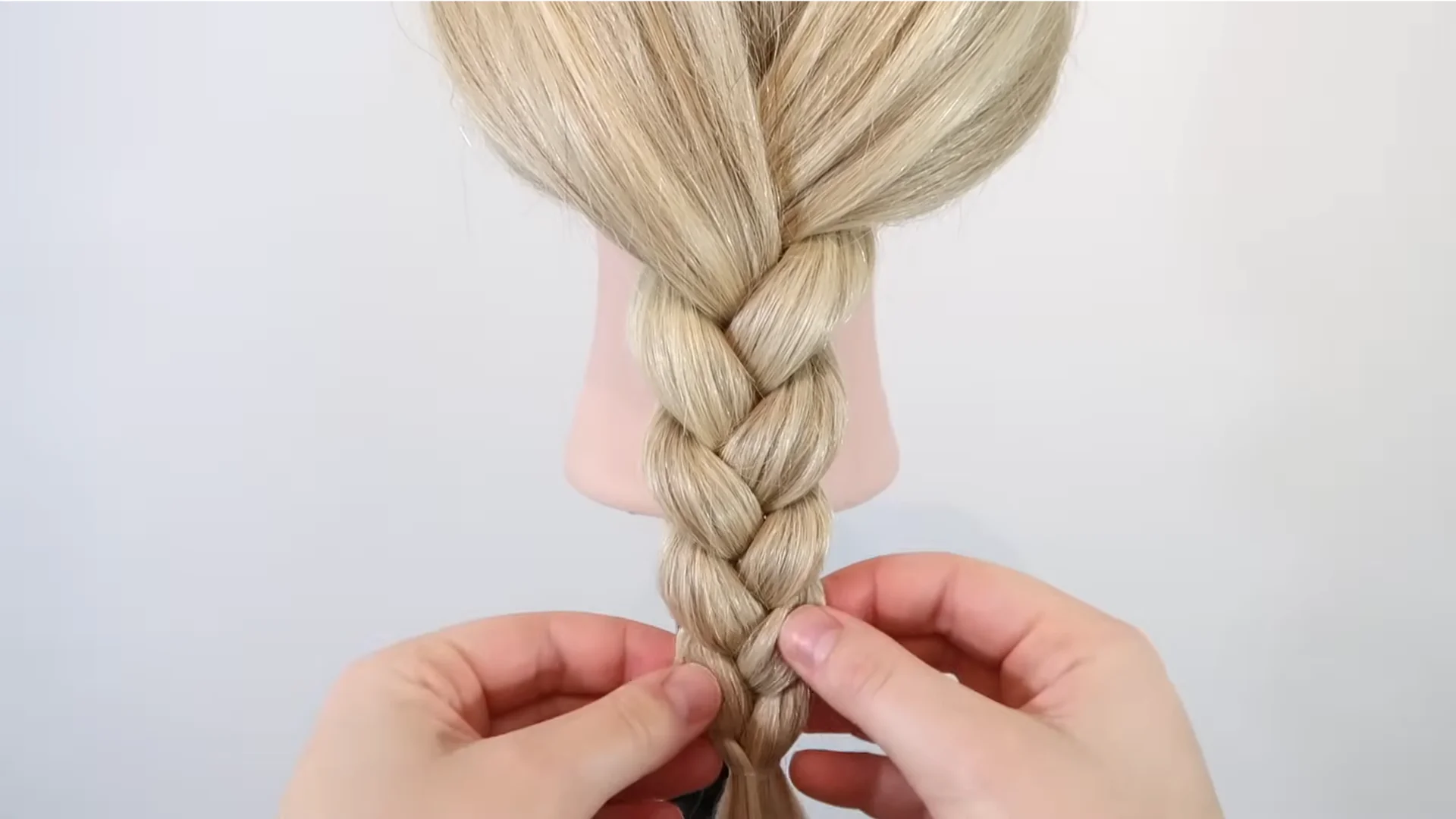 Hands gently pulling the sides of a finished blonde braid to loosen the woven sections and create a fuller, more voluminous look.