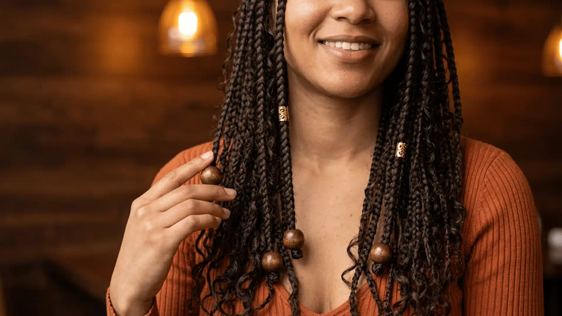 Light-skinned Black woman with boho braids adorned with wooden beads and gold cuffs against a dark wood wall