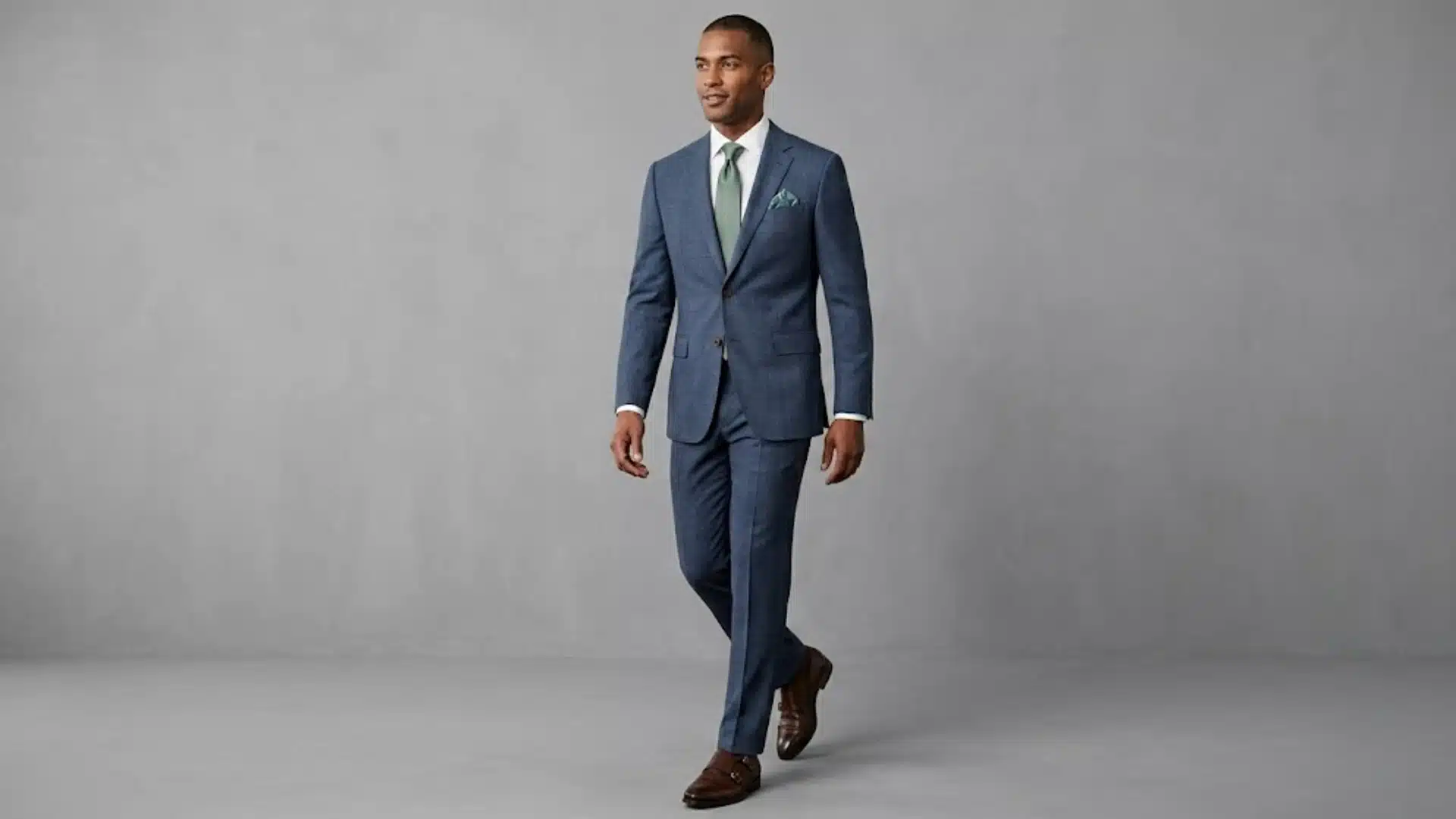 Man wearing a blue suit jacket with matching trousers, green tie, pocket square, and brown dress shoes