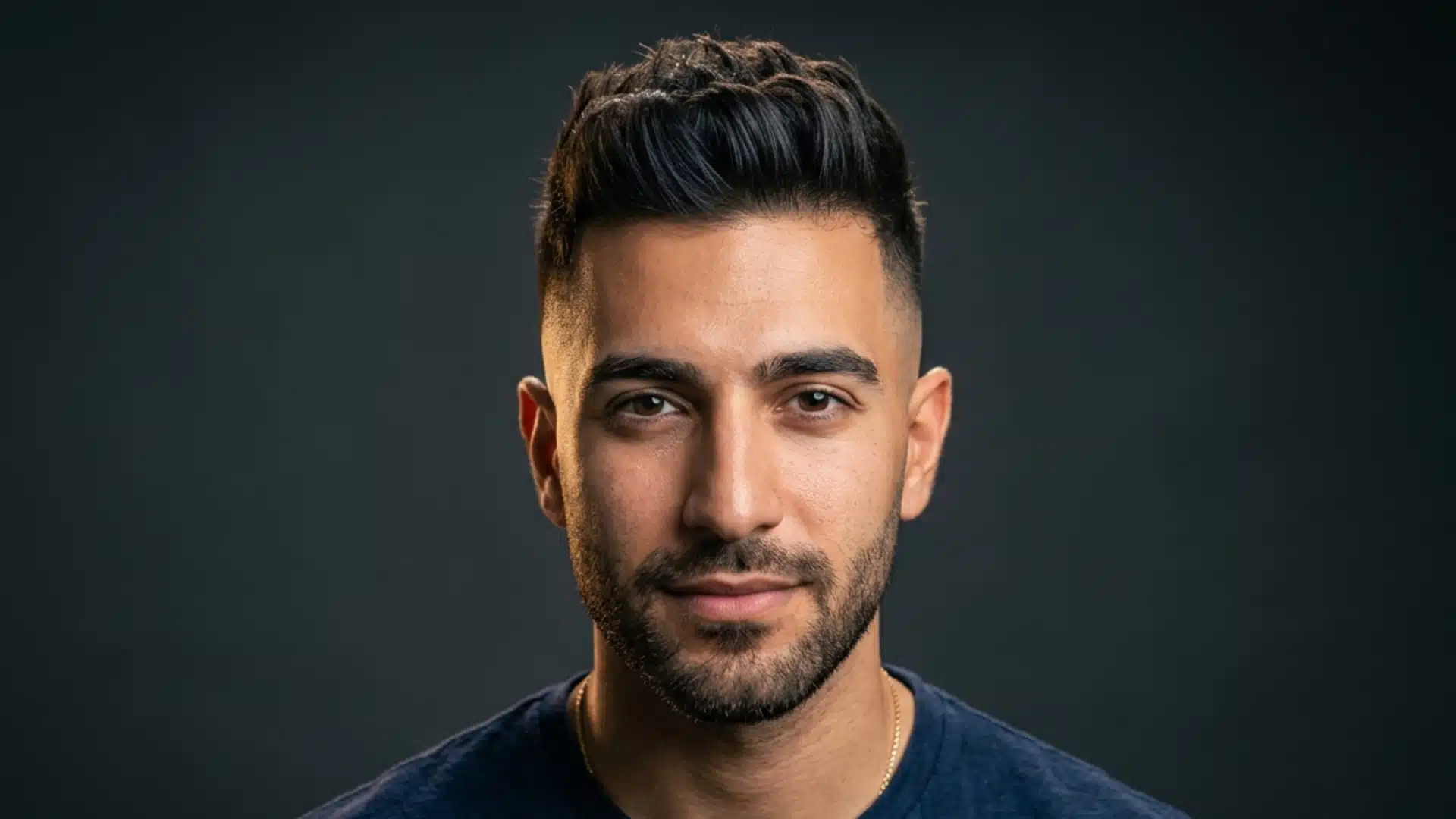 Man with a styled short quiff haircut and a beard looking forward against a dark background