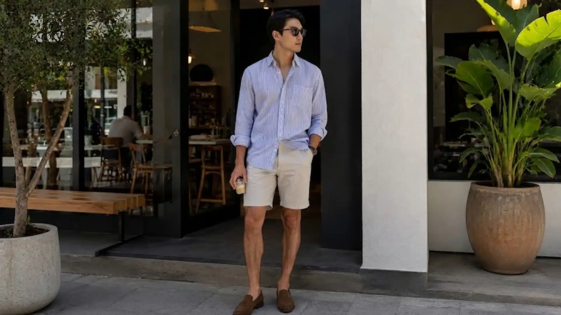 Man in a striped shirt, shorts, and sunglasses standing outside a cafe holding a drink