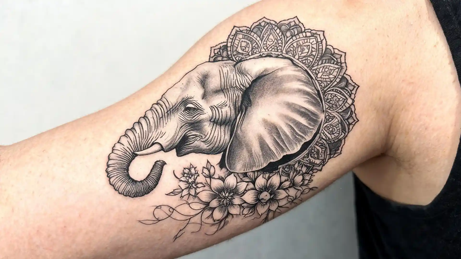 Realistic elephant upper arm tattoo with mandala halo and floral details in black and grey ink