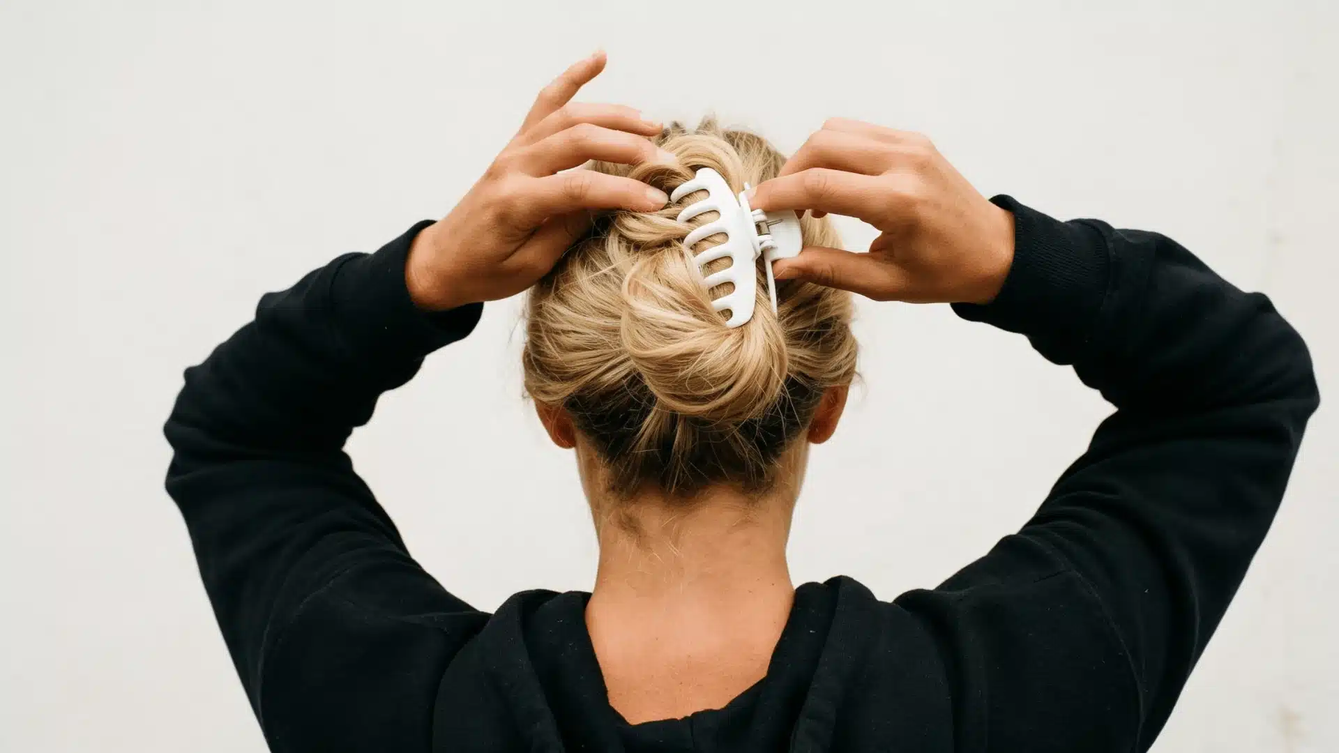 Rear view of a blonde woman wearing a black hoodie as she styles her hair upward using a white claw clip against a plain light wall